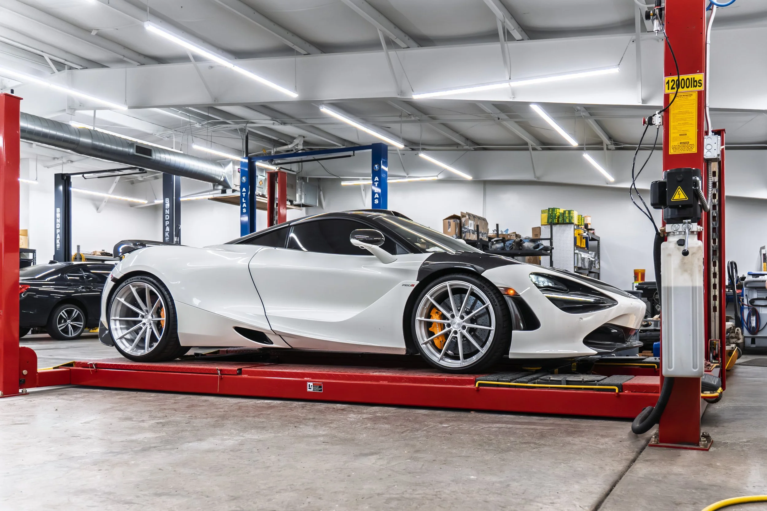 A white sports car on a red hydraulic lift inside an auto repair shop. The shop has bright lights, white walls, and various tools and equipment visible in the background.