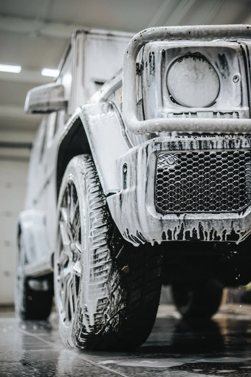 Front of a white vehicle being washed with soap foam in an indoor facility.