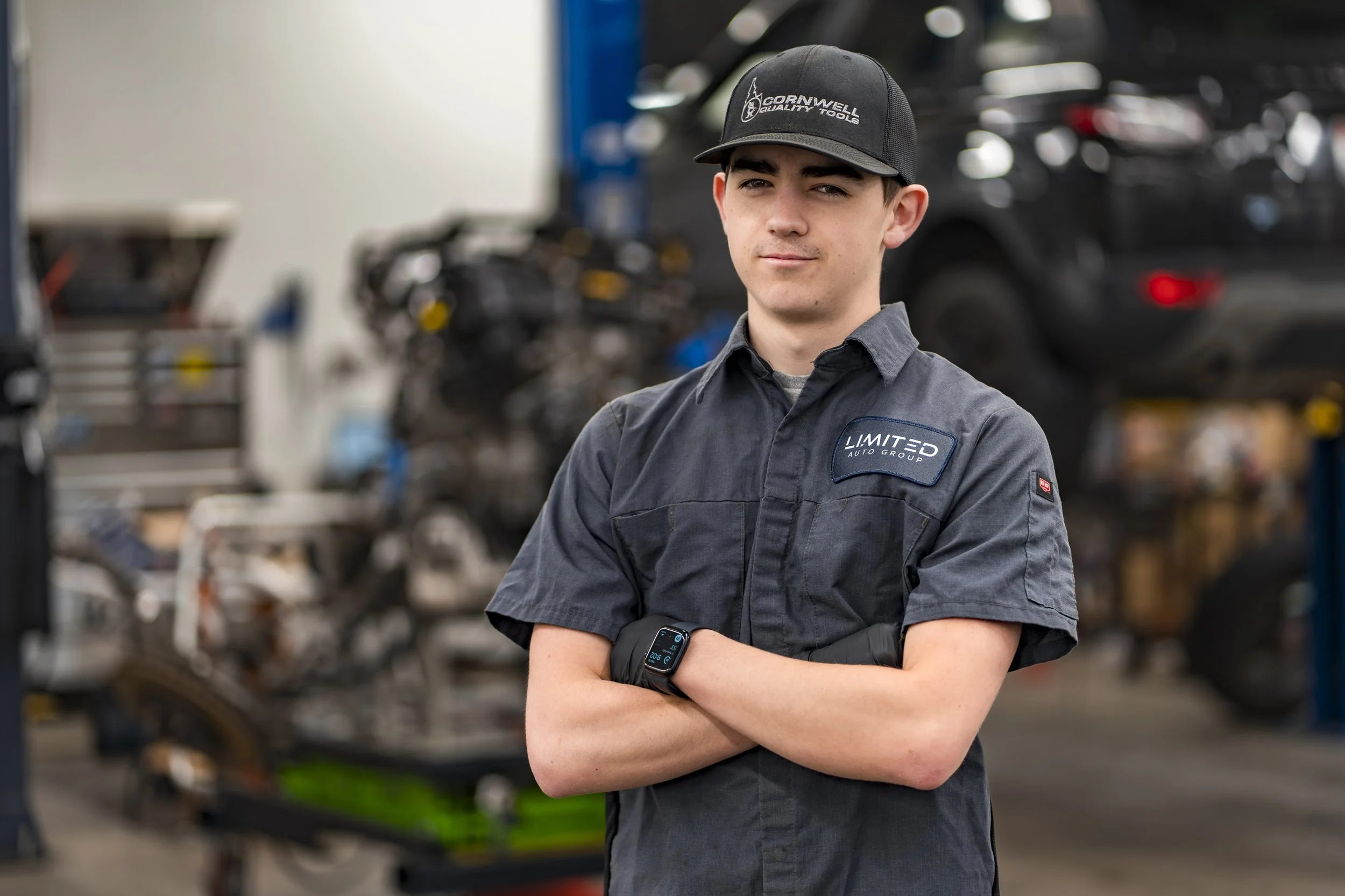 A young male mechanic with crossed arms wearing a dark uniform and a cap with the text "Cornwells Quality Tools" in an auto repair shop.