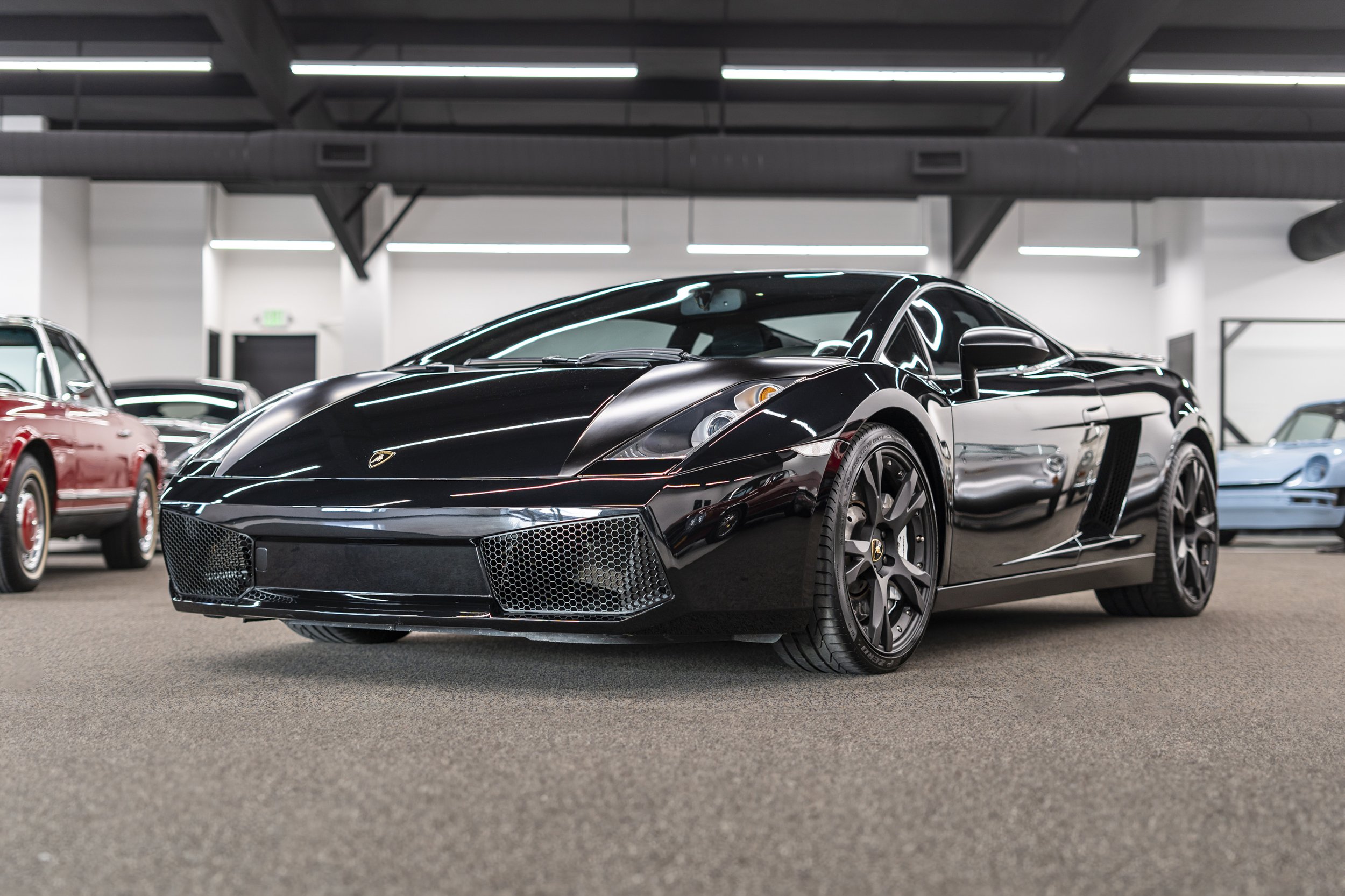 Black Lamborghini sports car inside a showroom with other luxury cars and ceiling lights reflected on its surface.