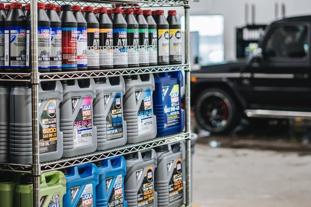 Shelves stocked with various bottles and containers of motor oil and automotive fluids in a car repair shop, with a black vehicle in the background.
