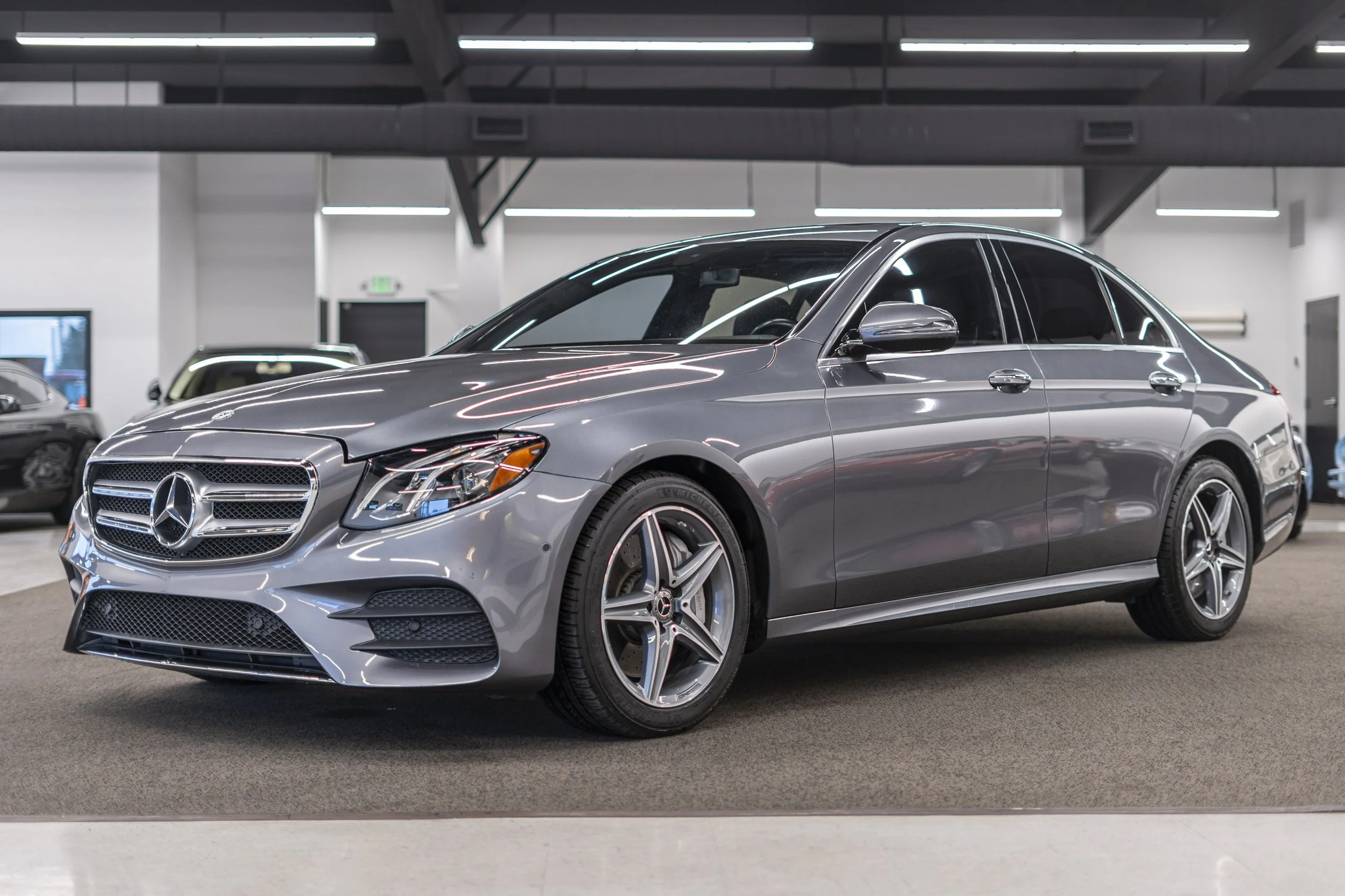 A silver Mercedes-Benz sedan parked indoors in a showroom or garage with ceiling lights, other cars, and a door visible in the background.
