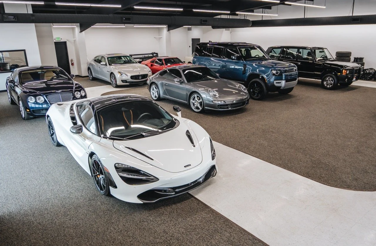Luxury cars parked indoors, including a white McLaren, black Bentley, silver Maserati, red sports car, gray Porsche, blue Range Rover, and black classic Land Rover with tires stacked in the background.