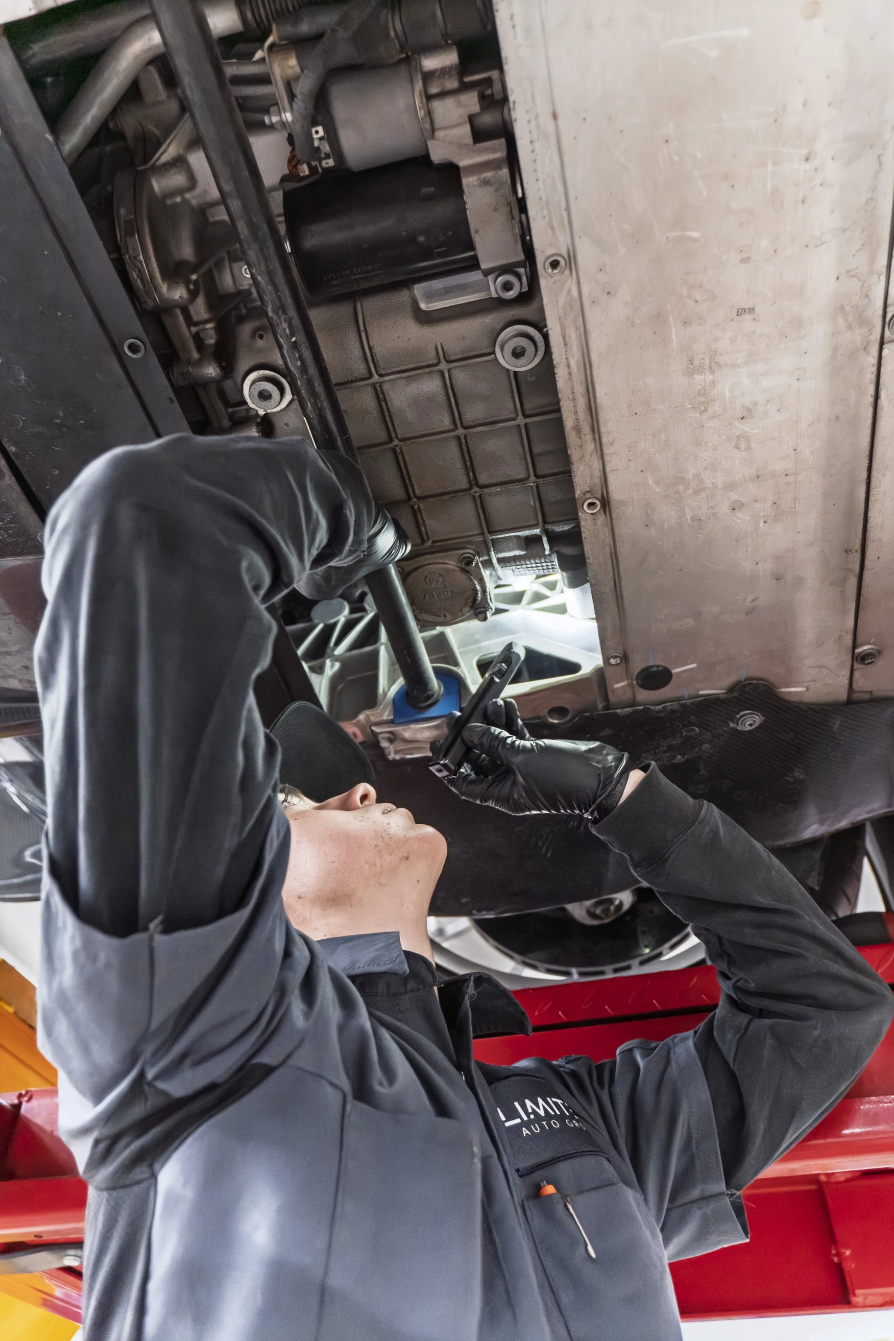 A mechanic working underneath a vehicle, wearing black gloves and a gray uniform, with a red mechanic lift visible in the background.