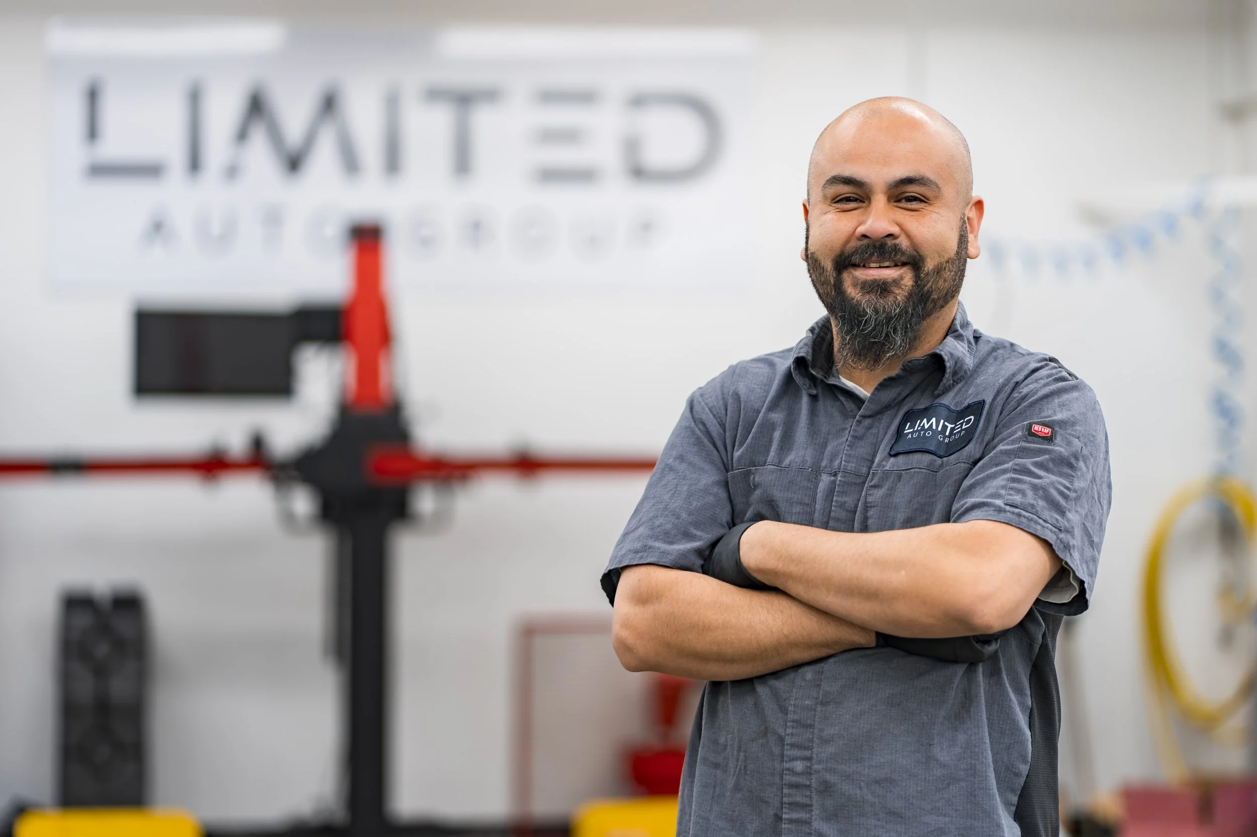 A smiling man with a beard and shaved head stands with arms crossed in a workshop or garage with blurred equipment and a sign that says "Limited" in the background.