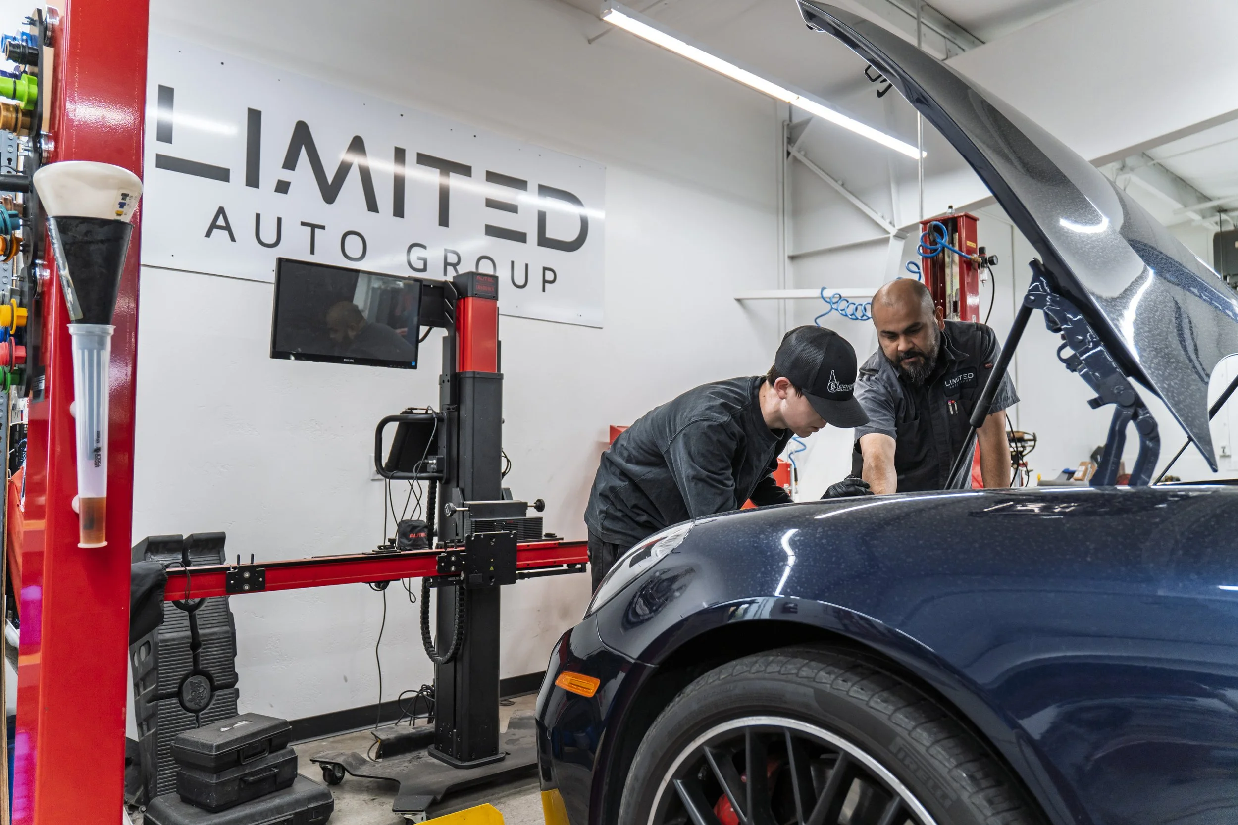 Two mechanics working on a black sports car in a garage. The garage has a large sign that reads 'LIMITED AUTO GROUP'. Mechanical tools and equipment are visible around them.