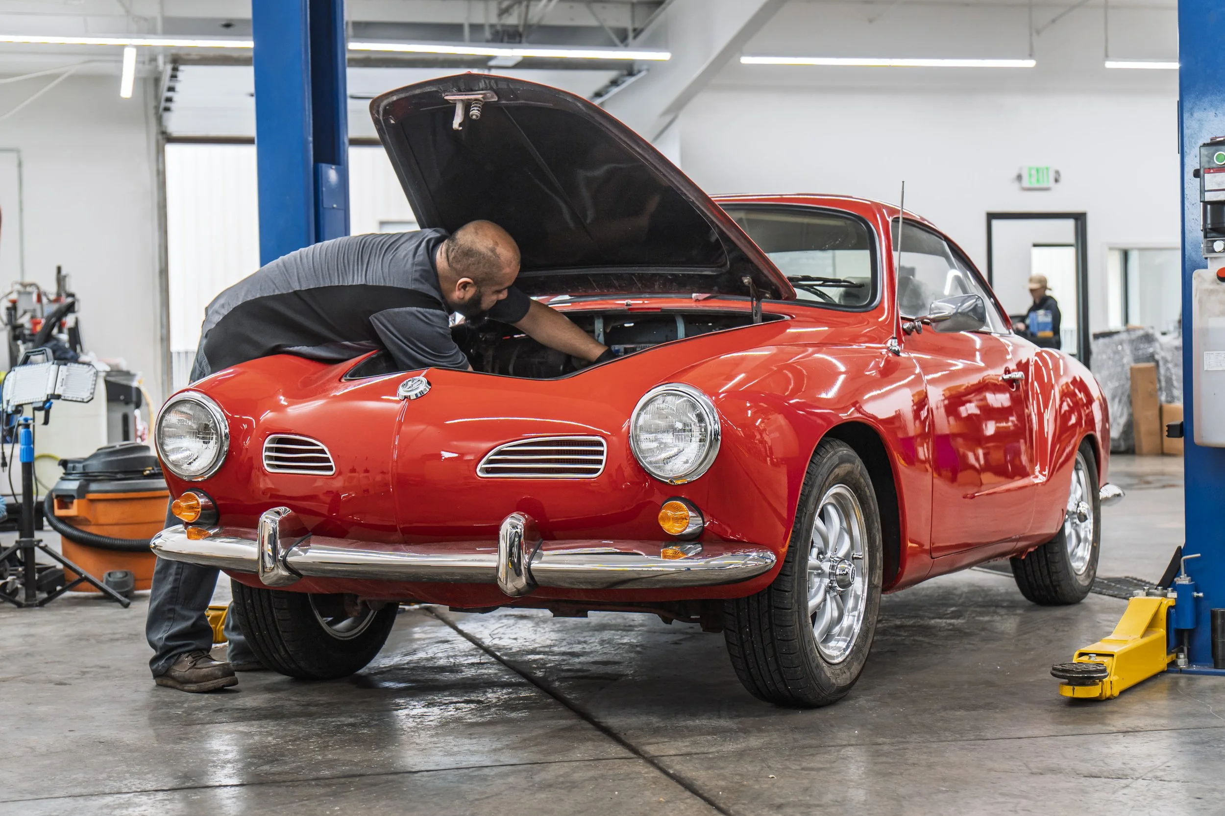 Man working on a vintage red sports car inside an auto workshop, inspecting under the hood.