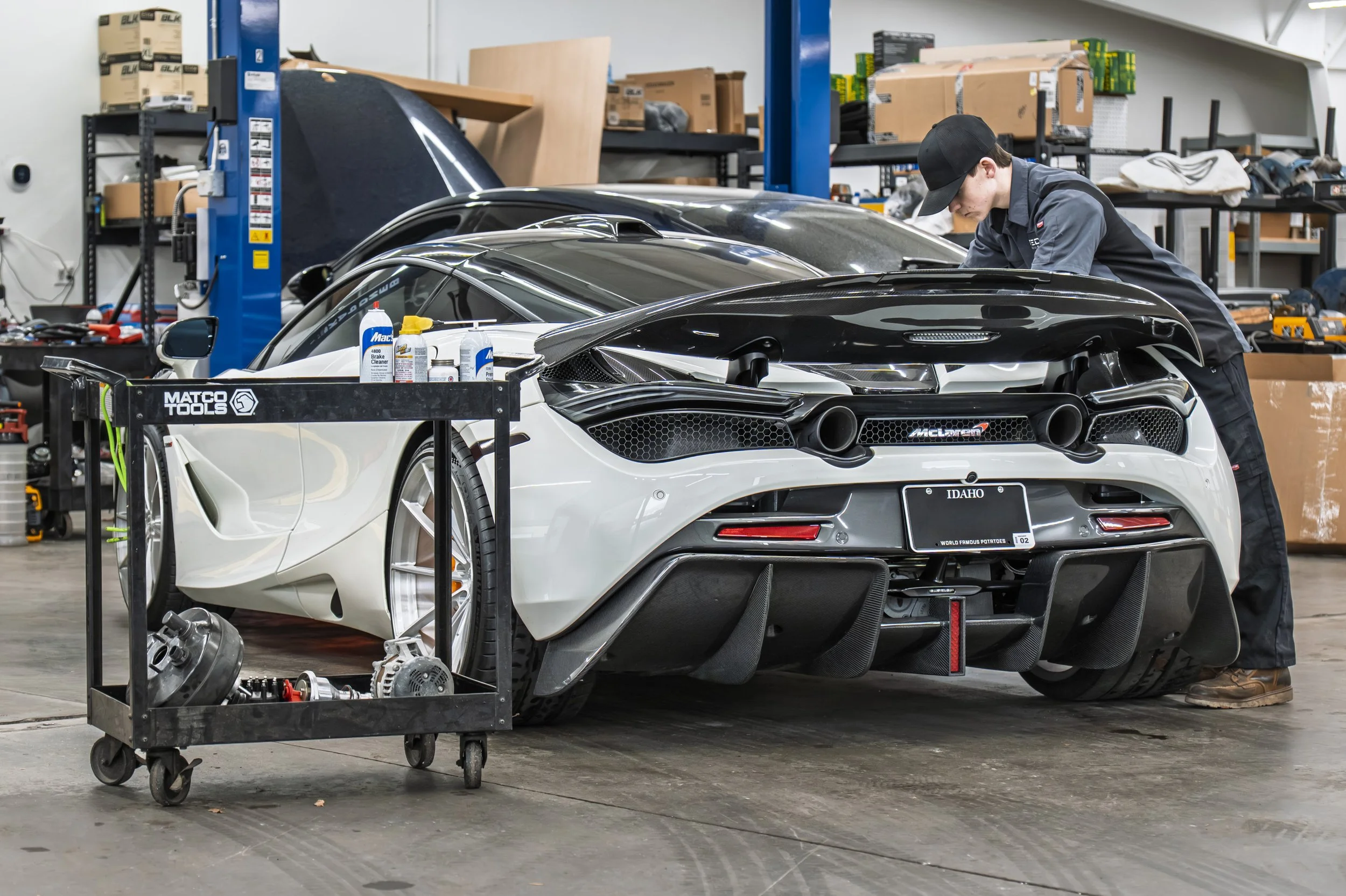A mechanic working on a white McLaren sports car inside an auto workshop. Tools and car parts are on a cart in the foreground. Shelves with boxes and supplies are in the background.