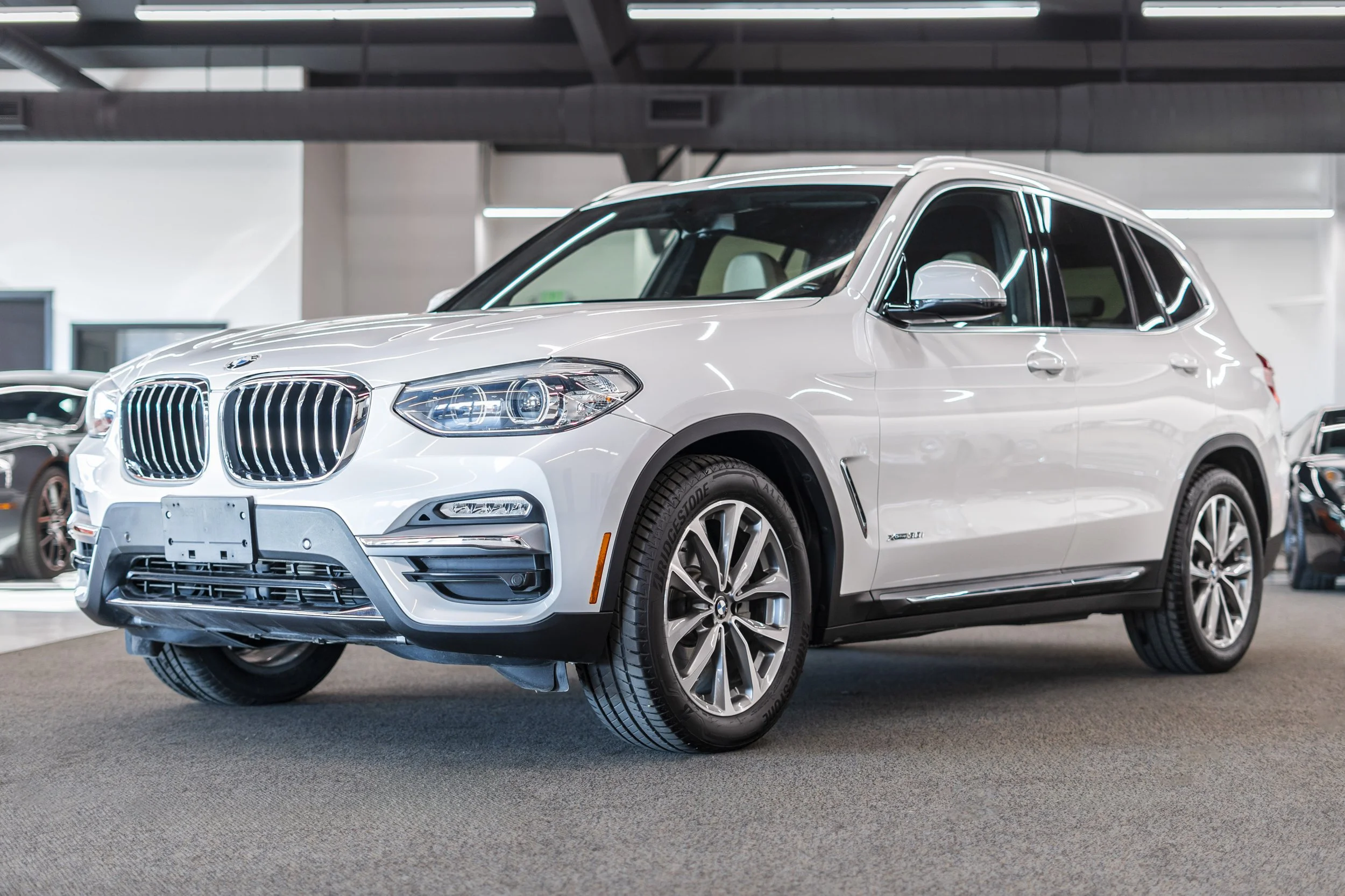 A white BMW SUV parked indoors, likely in a dealership showroom, with bright lighting reflecting off its surface.