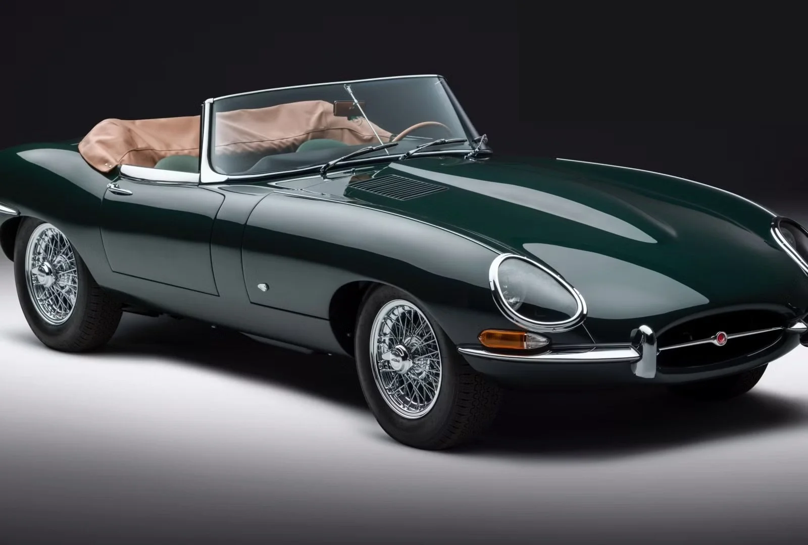 A classic dark green Jaguar E-Type convertible sports car with wire wheels and tan interior, set against a black background.