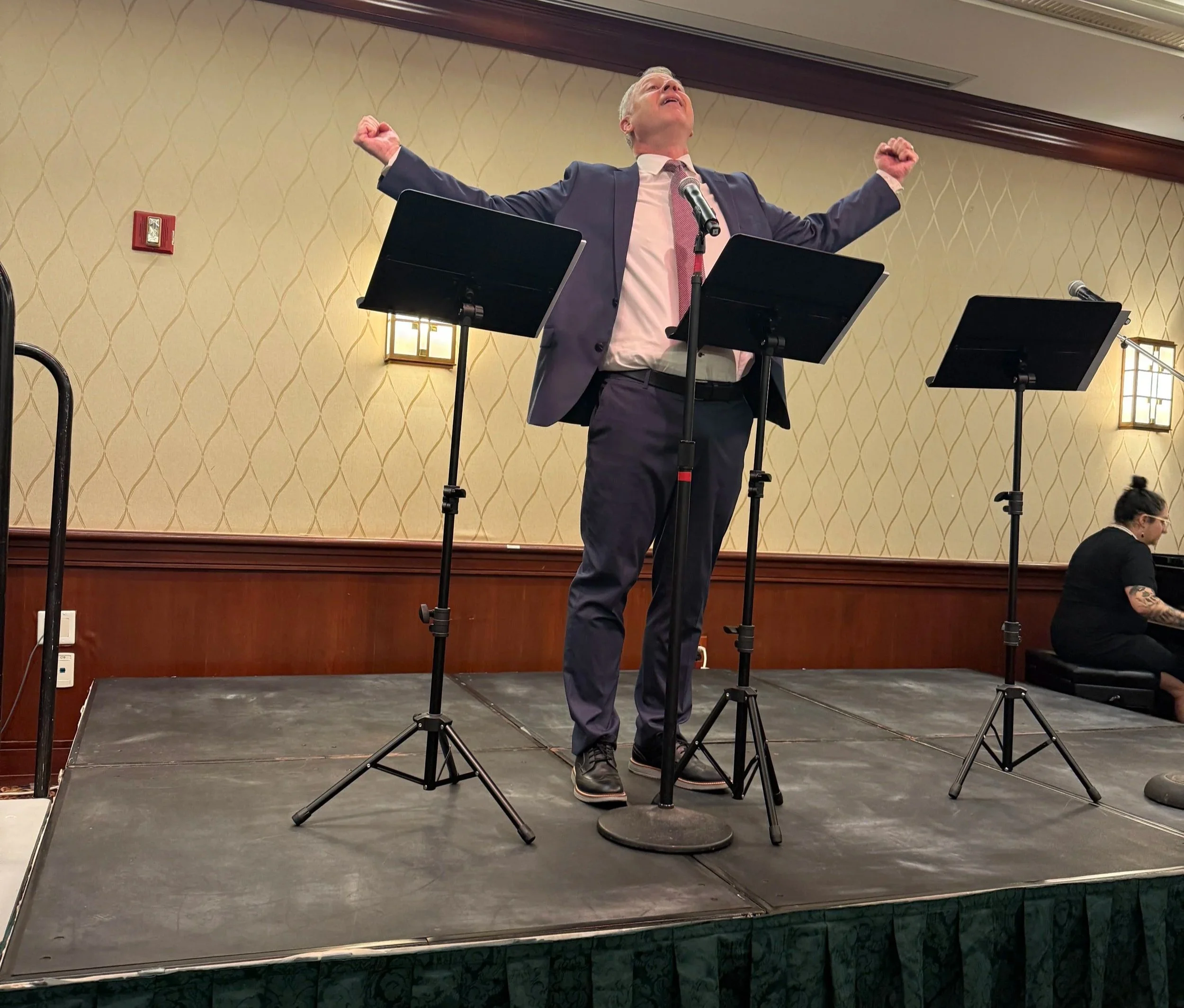A man in a suit stands on a stage with his arms raised in victory, speaking into a microphone, with three music stands in front of him, in a conference room.