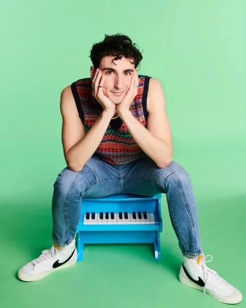 Young man sitting on a small blue piano bench, resting his chin on his hands, smiling at the camera, wearing a sleeveless multicolored shirt, blue jeans, and white Nike sneakers with black swooshes, against a light green background.