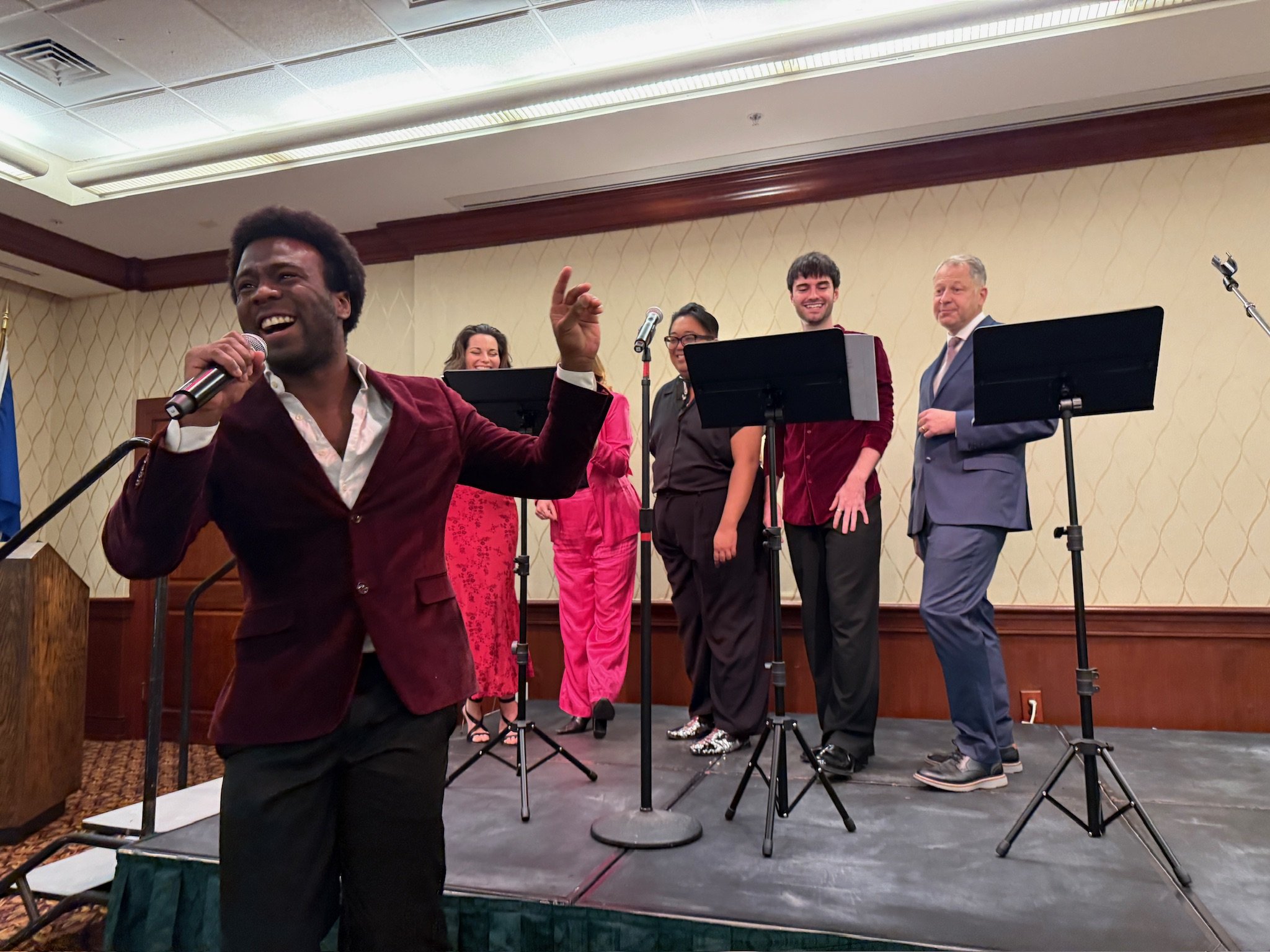 A group of six people on a stage at an indoor event. One man in a burgundy velvet blazer is holding a microphone and smiling, while the other five are standing behind music stands, laughing and engaging with the audience.