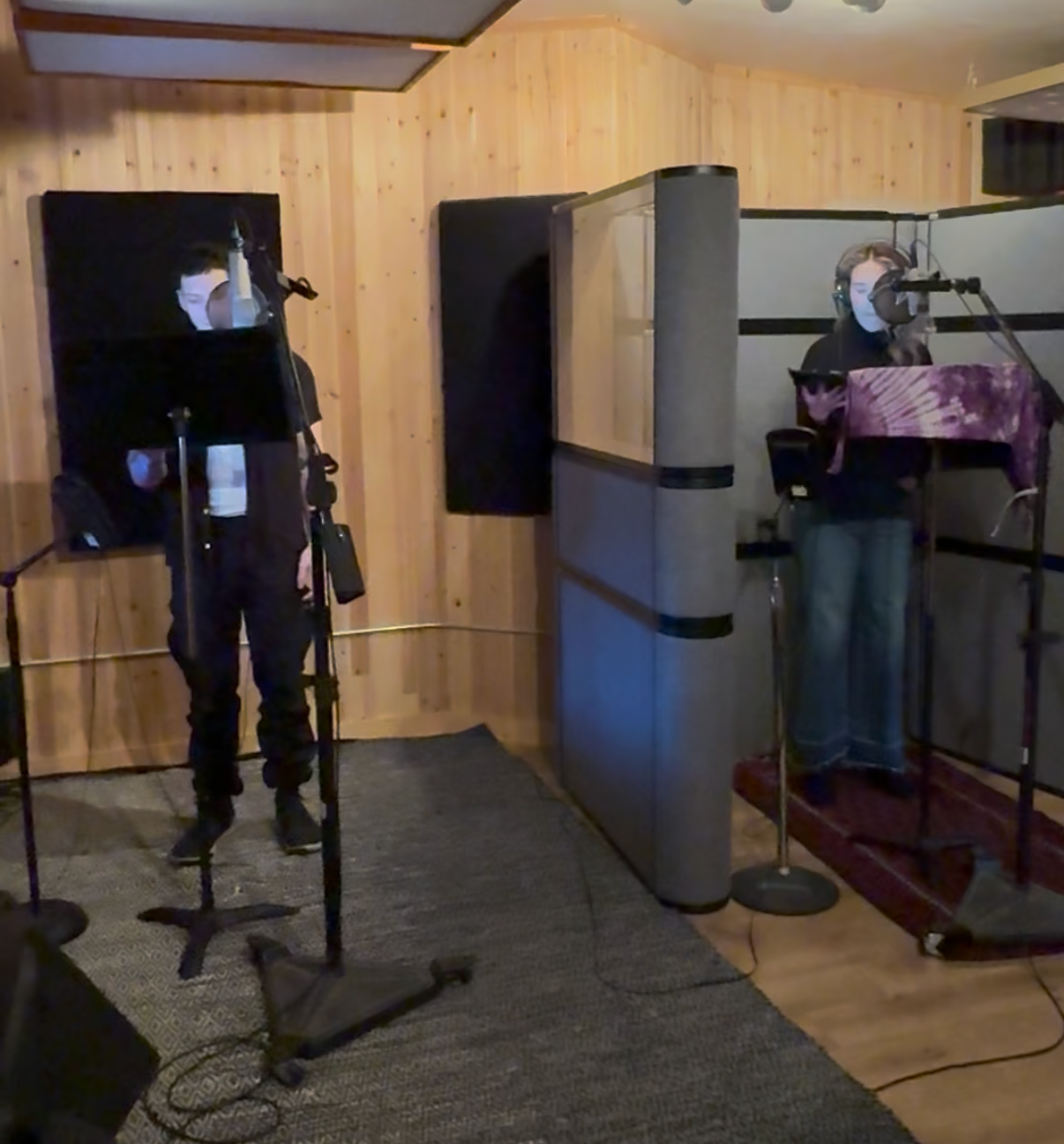 Two people in a recording studio wearing headphones and face masks, standing behind microphone and music stands, surrounded by soundproofing panels and wooden walls.
