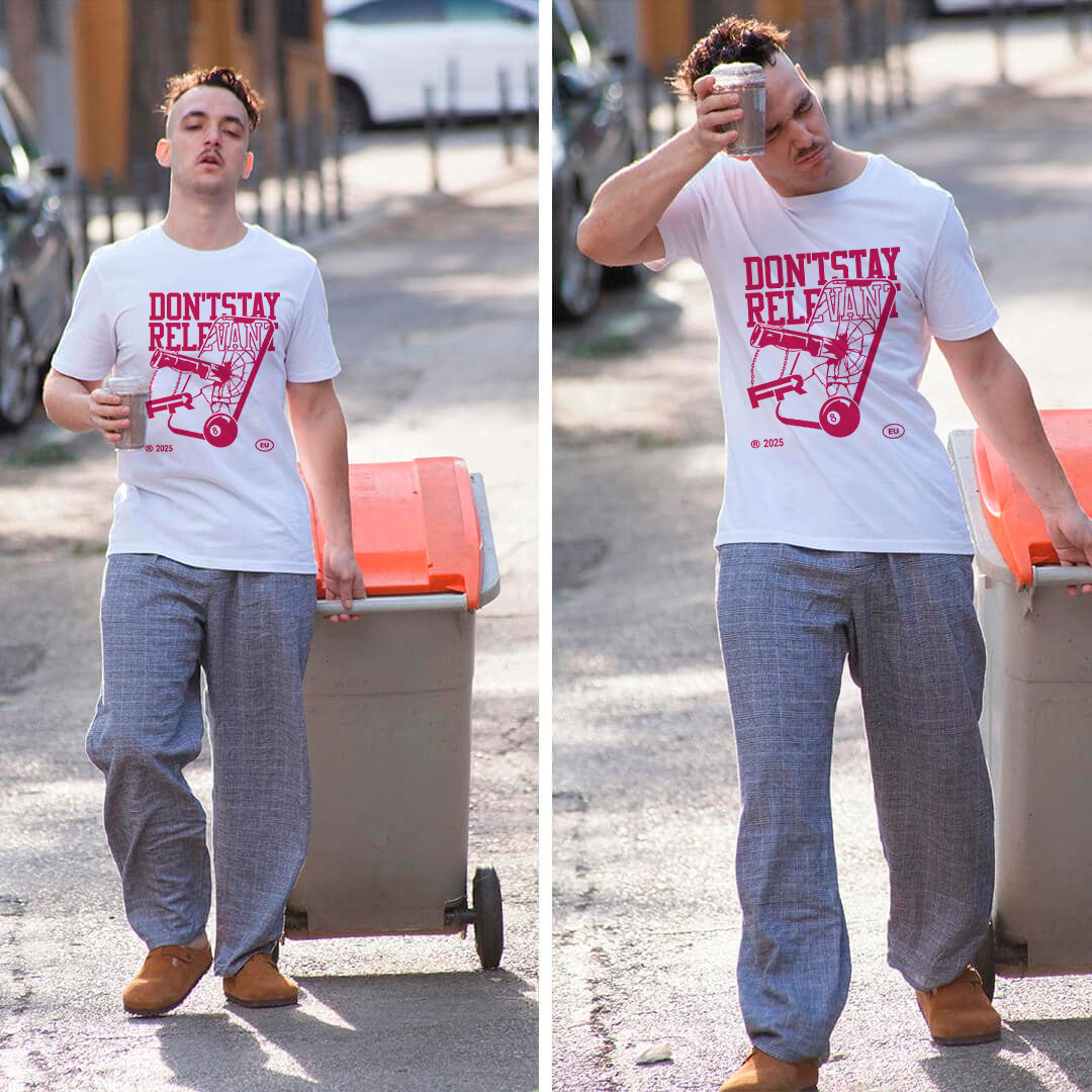 Hombre joven caminando con un carrito de basura, sosteniendo un vaso con bebida, vistiendo camiseta blanca con texto 'Don't Stay Relax', pantalones gruesos y botas de color marrón.