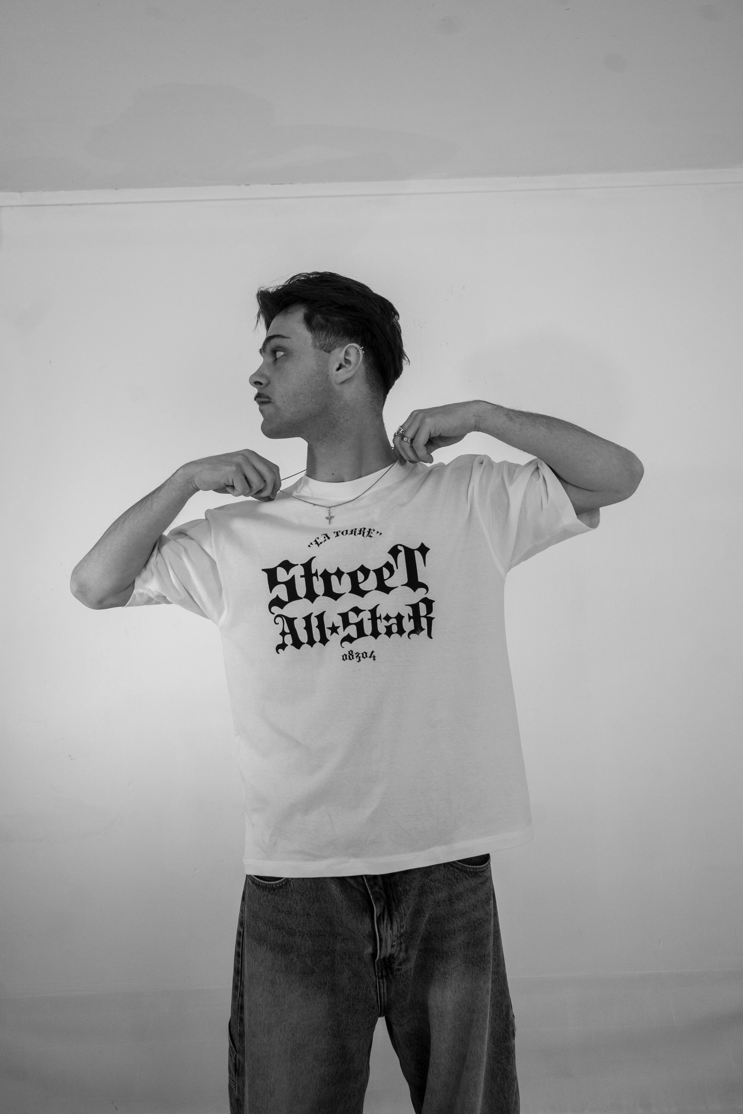 Joven hombre con camiseta blanca que dice 'Street All-Stark' y pantalones vaqueros, ajustándose el collar.