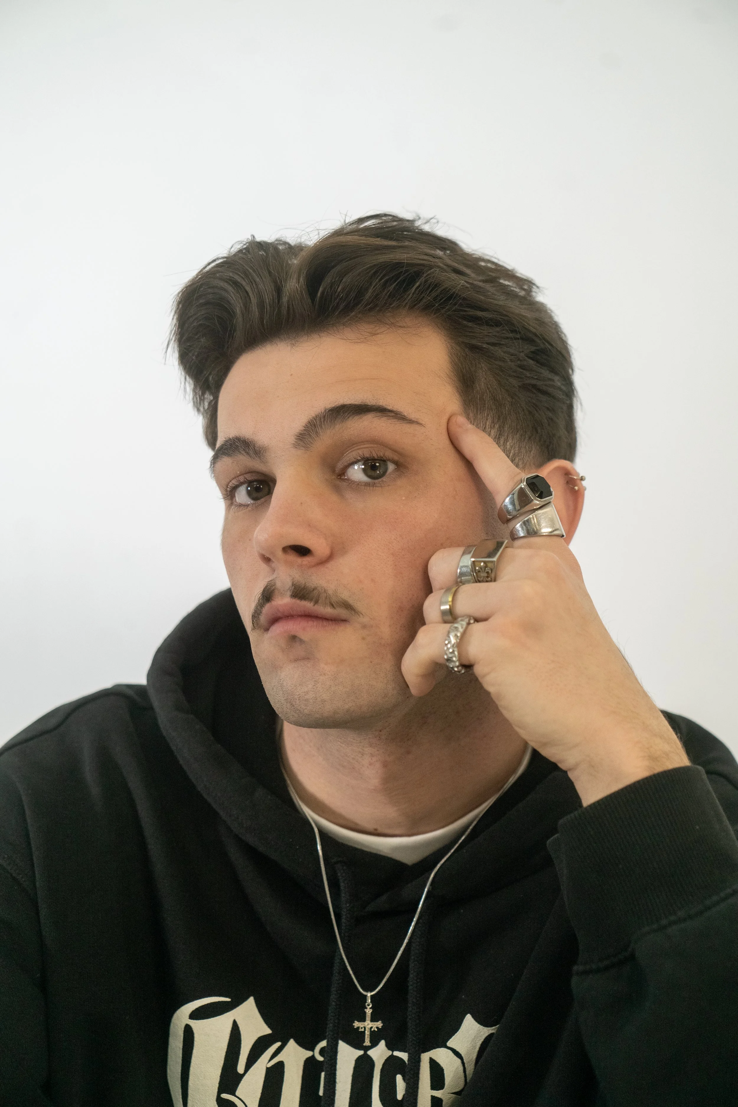 Joven con estilo punk, con múltiples anillos y piercings, posando con expresión pensativa en fondo blanco.