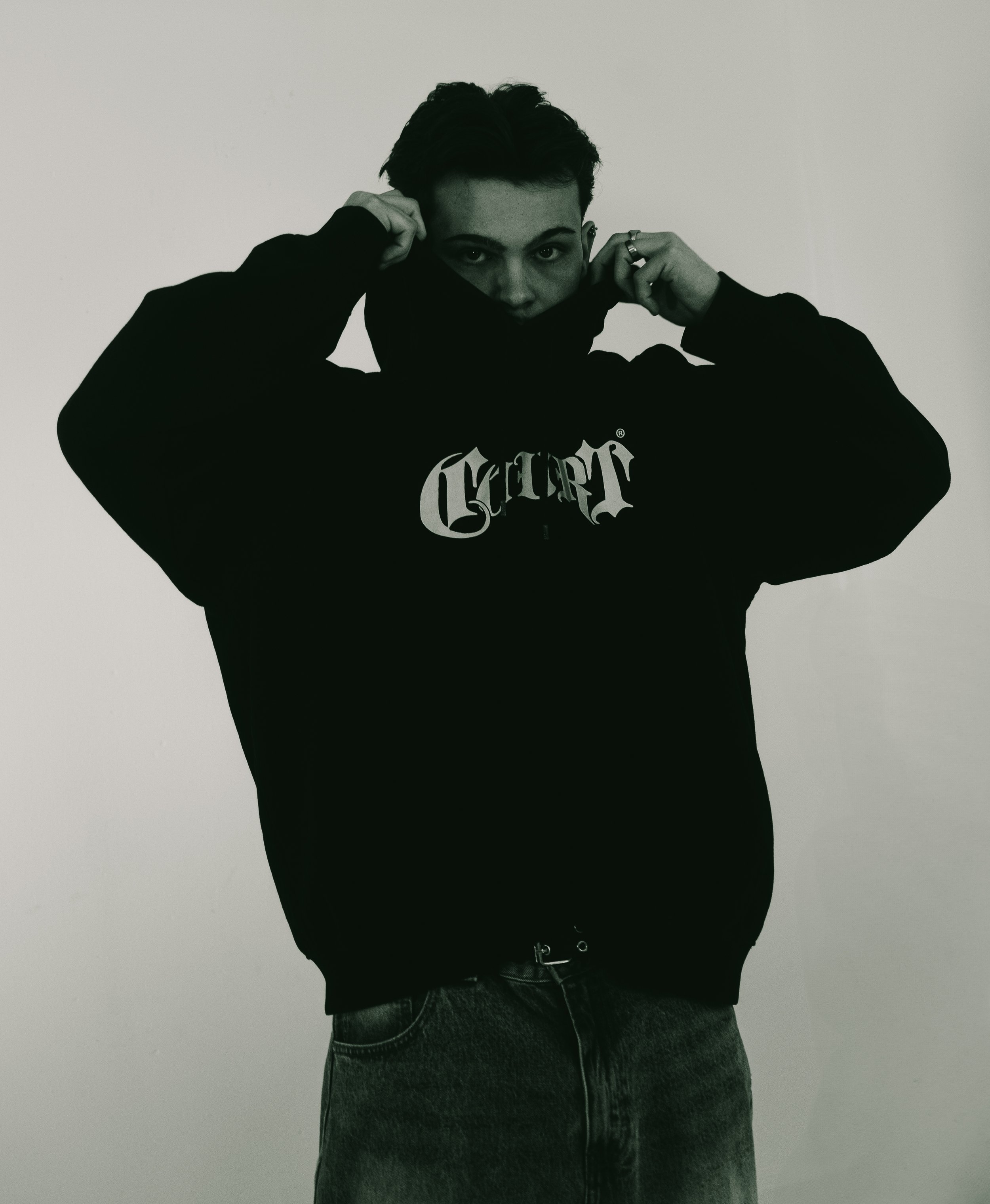 Joven con sudadera negra y pantalones vaqueros, cubriéndose parcialmente la cara, en un fondo neutral.