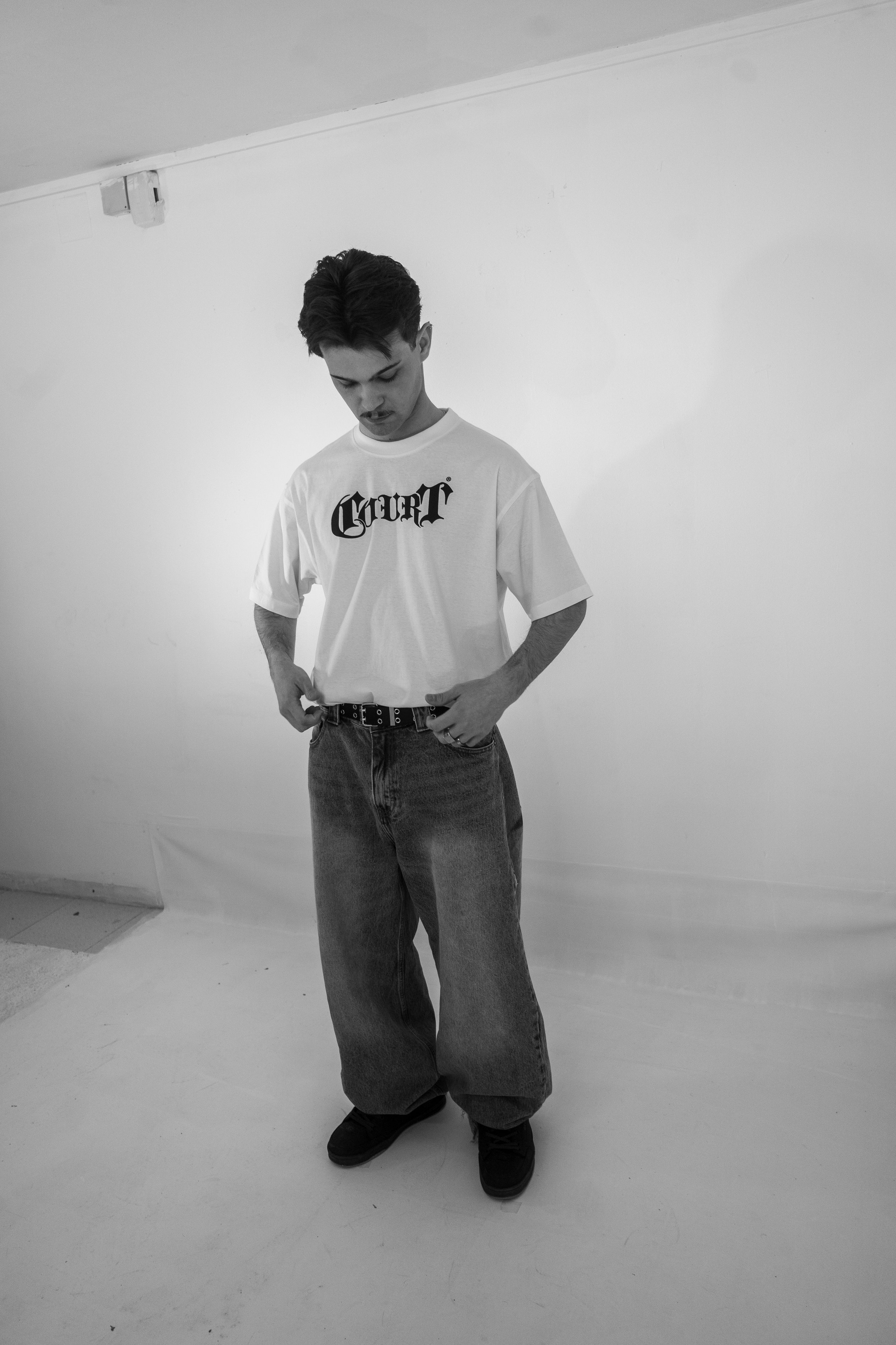 Un joven de cabello oscuro, camiseta blanca con la palabra 'Curry' en letras negras y pantalones anchos de mezclilla, está parado en una pared blanca en un estudio fotográfico.