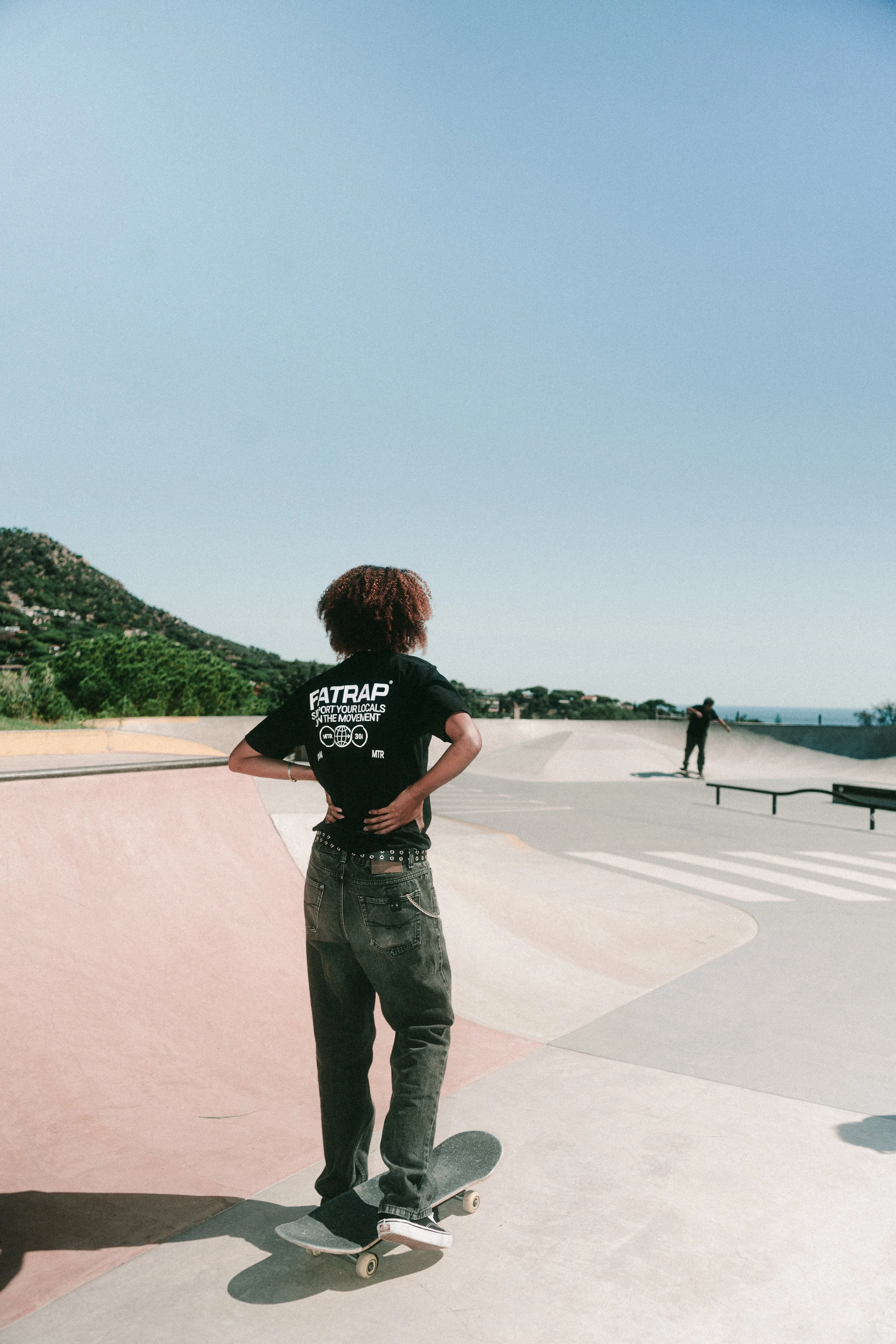 Una joven en patineta en un skatepark al aire libre, con una camiseta negra que dice 'FATRAP' y pantalones oscuros, en un día soleado con cielo despejado.
