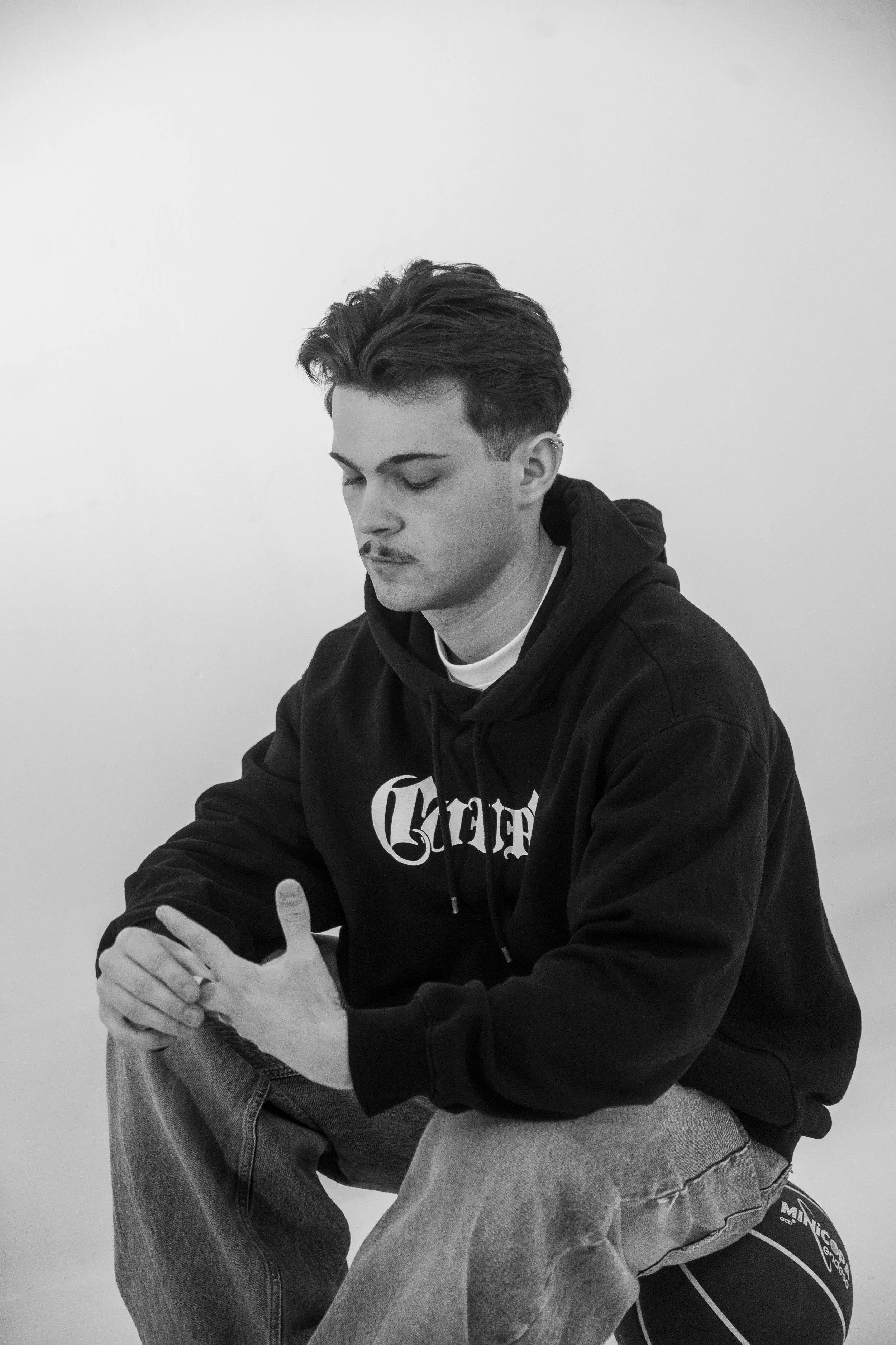 Un joven masculino con cabello oscuro y peinado hacia un lado, usa una sudadera negra y jeans, sentado con las manos juntas y mirando hacia abajo, en un fondo blanco y sin otros elementos visibles.