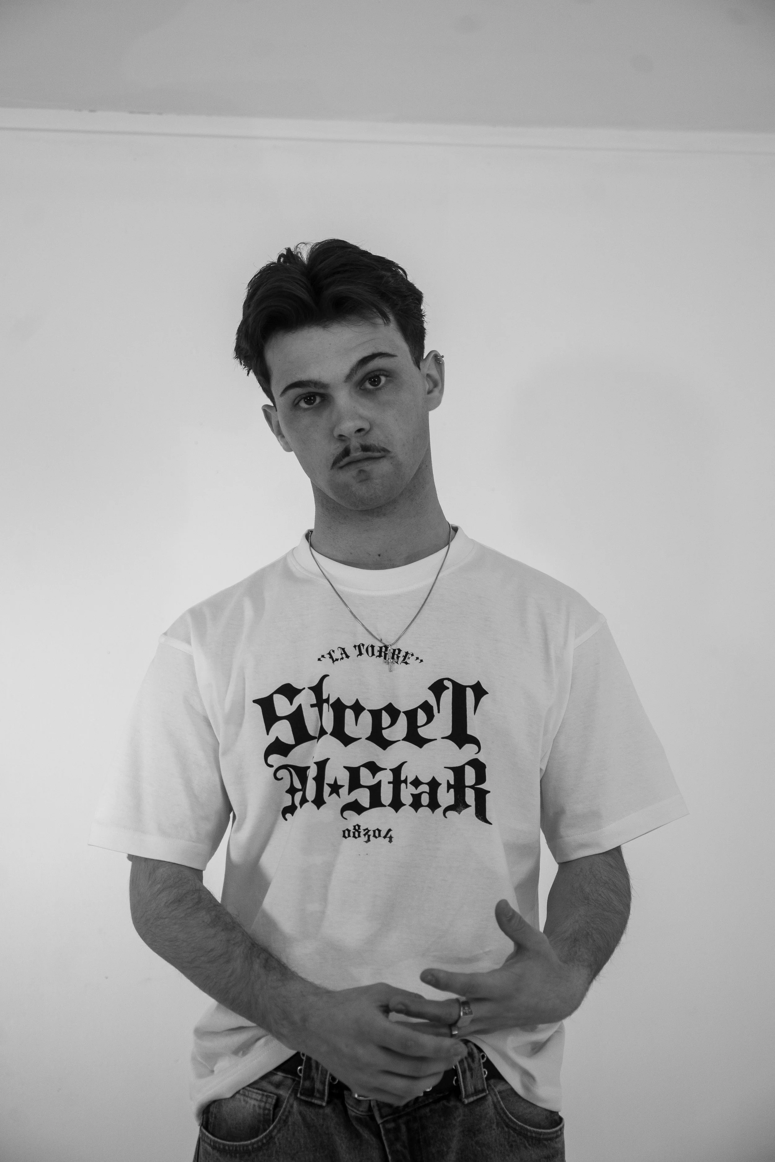 Un joven con peinado oscuro y barba delgada, vistiendo una camiseta blanca con letras negras y un collar, posa frente a una pared blanca en un entorno sencillo en blanco y negro.