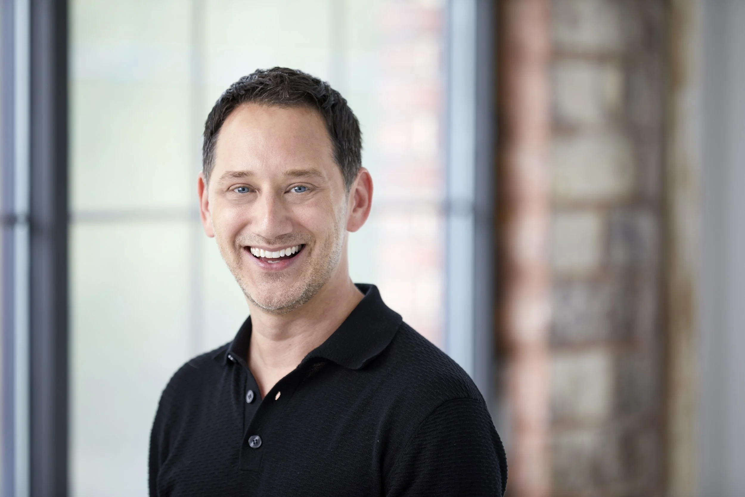 A photo of Jeffrey from B&Co. wearing a navy blue zip-up sweater over a white collared shirt, standing by a doorway with a conference room in the background.