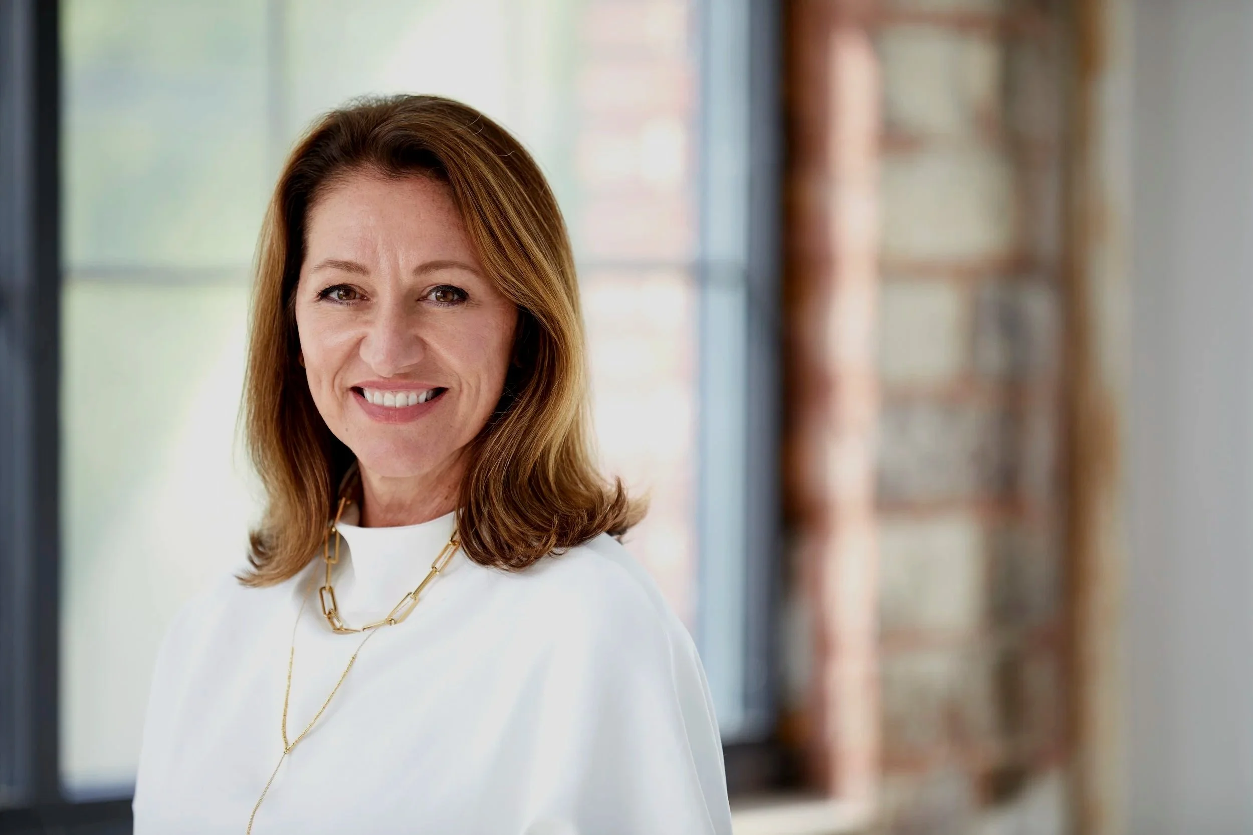 A photo of Brigette from B&Co. wearing a white top and gold jewelry, smiling in a bright indoor setting with windows and brick walls in the background.