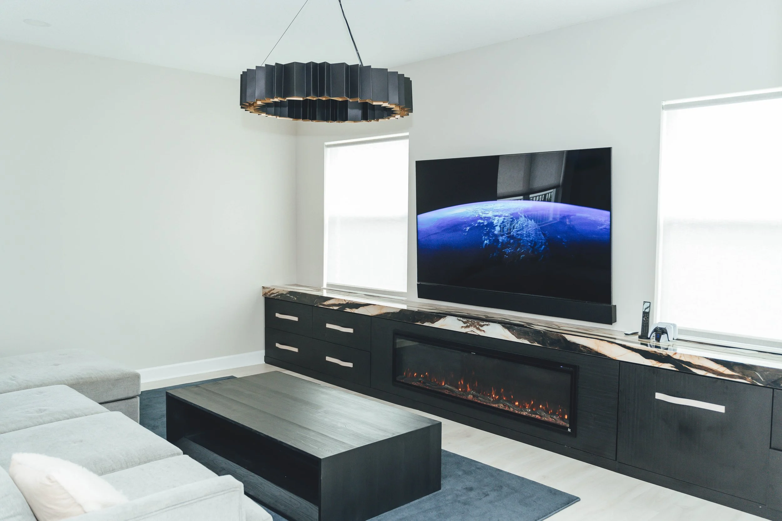 Modern living room with a black and gold chandelier, large flat-screen TV displaying Earth from space, a black fireplace, and a marble-topped console table along the wall. There are two windows with white blinds, a gray sofa, and a dark wood coffee t