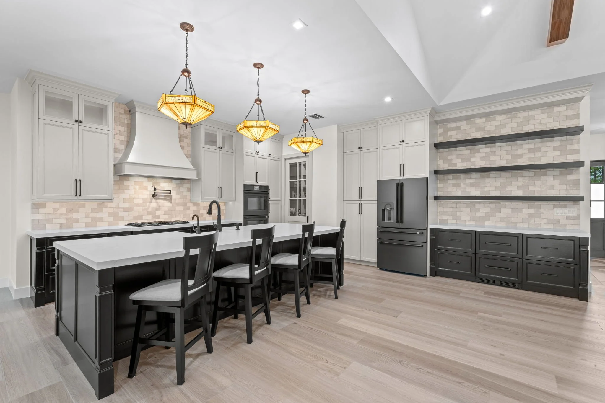 Modern kitchen with white cabinets, black appliances, a large kitchen island with black base and bar stools, beige brick backsplash, and three pendant lights.