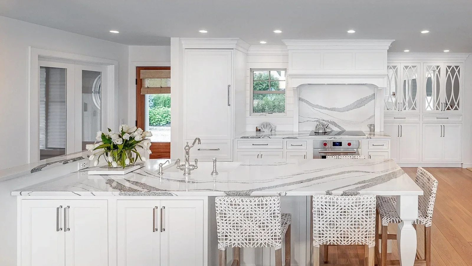 Modern white kitchen with marble countertops, centered on a kitchen island with a sink and decorative vase, and includes white cabinetry, a built-in oven, and wood accents.