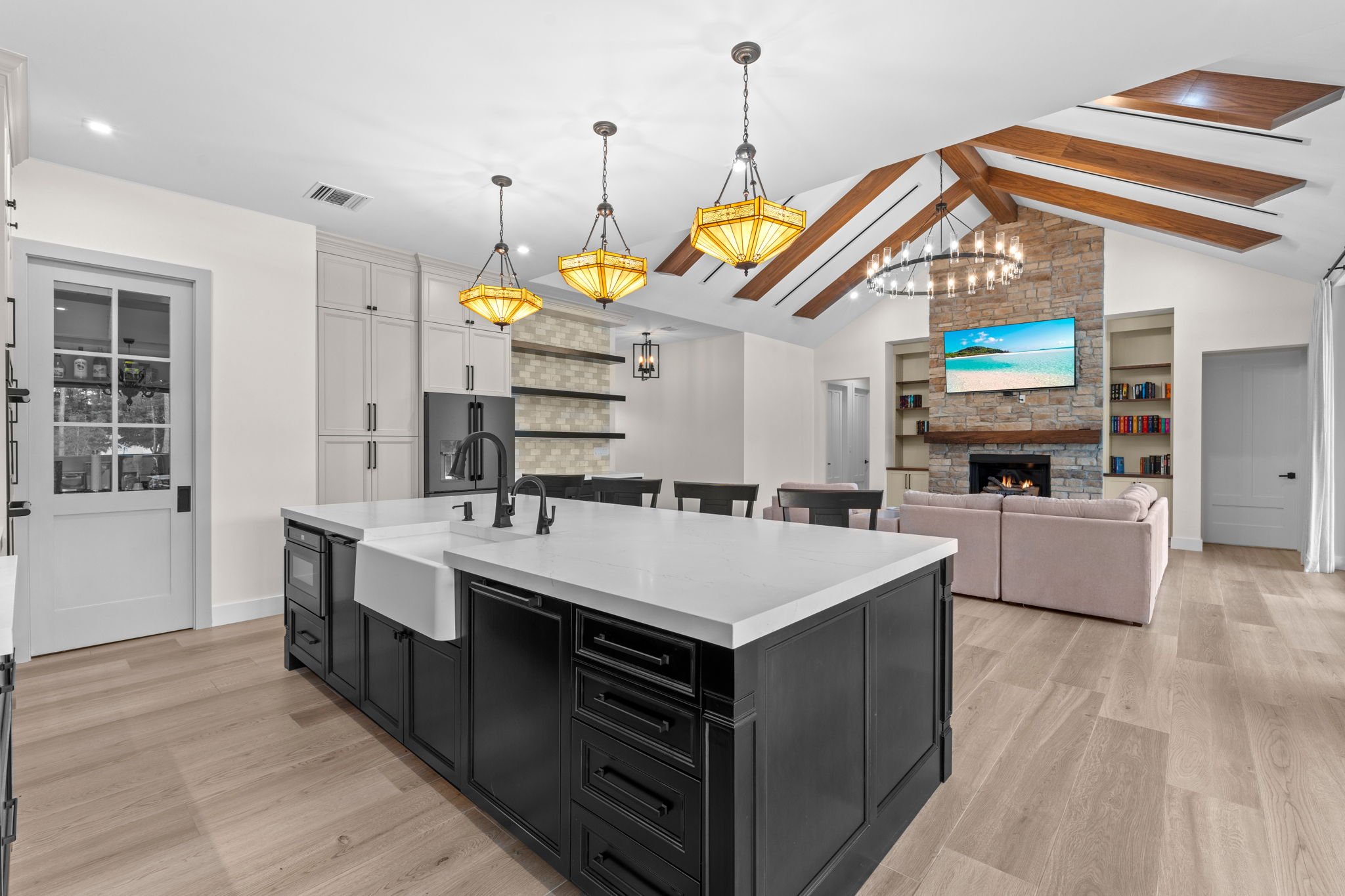Open-concept kitchen and living room with black kitchen island, white cabinetry, wooden ceiling beams, stone fireplace, flat-screen TV, and beige sectional sofa.