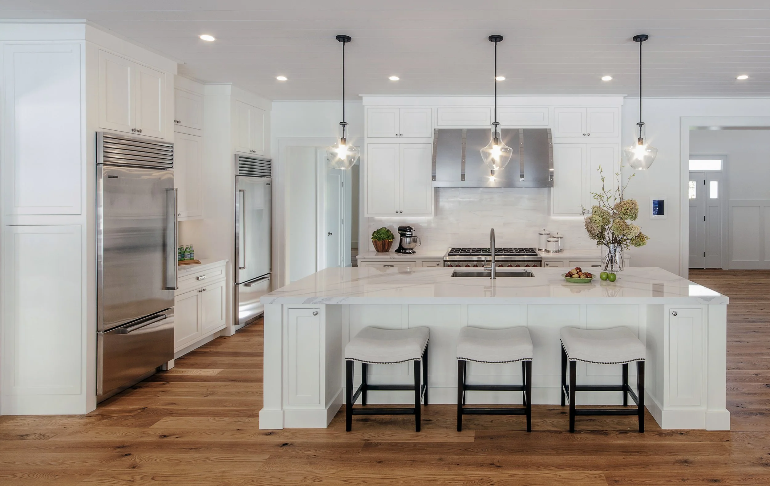 Modern white kitchen with wooden floor, large island with three white stools, four pendant lights, stainless steel appliances, flowers in a vase, and some fruit on the island.