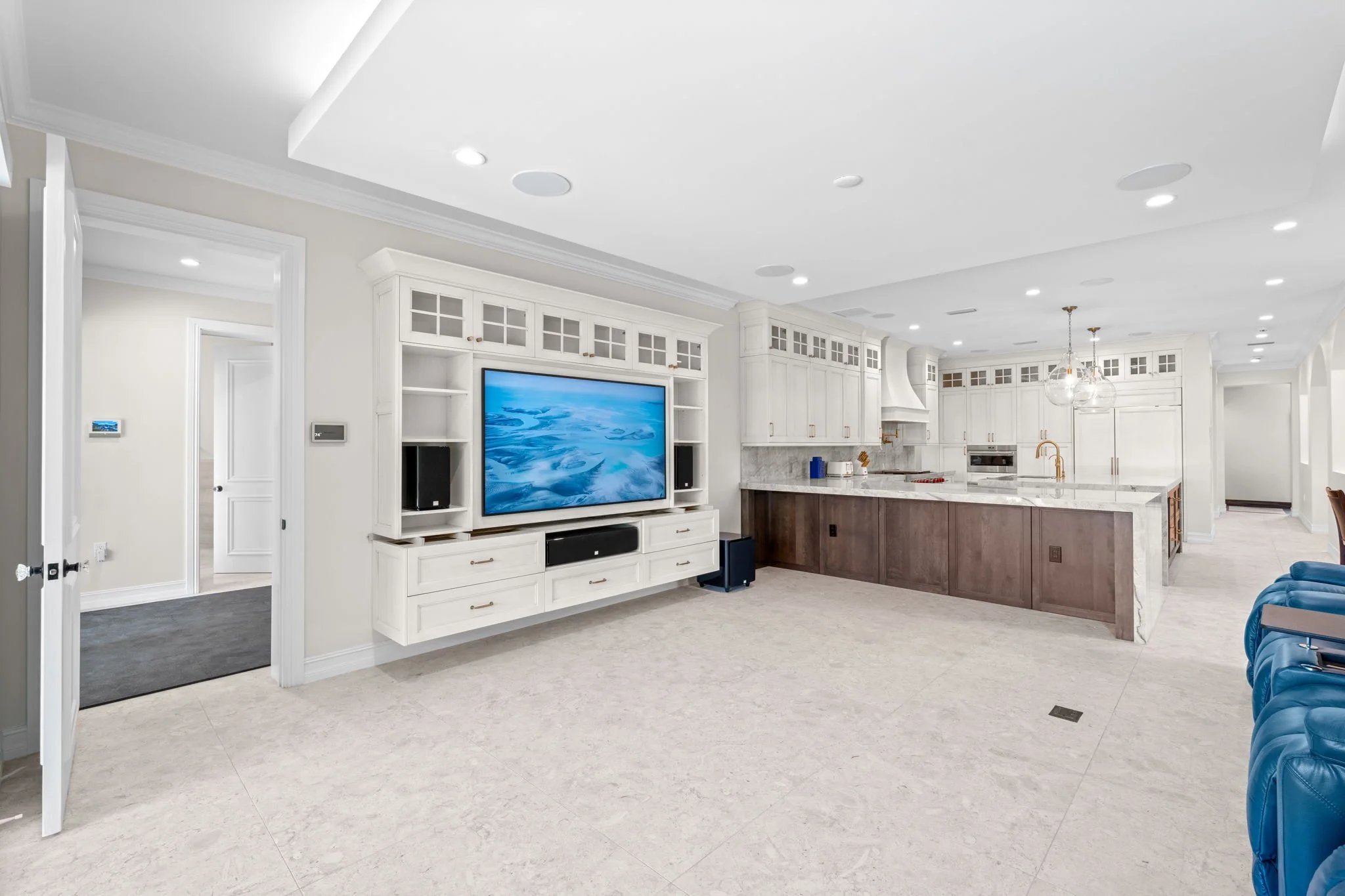 Bright, modern kitchen and living area with white cabinetry, marble countertops, a large flat-screen TV, and blue chairs.