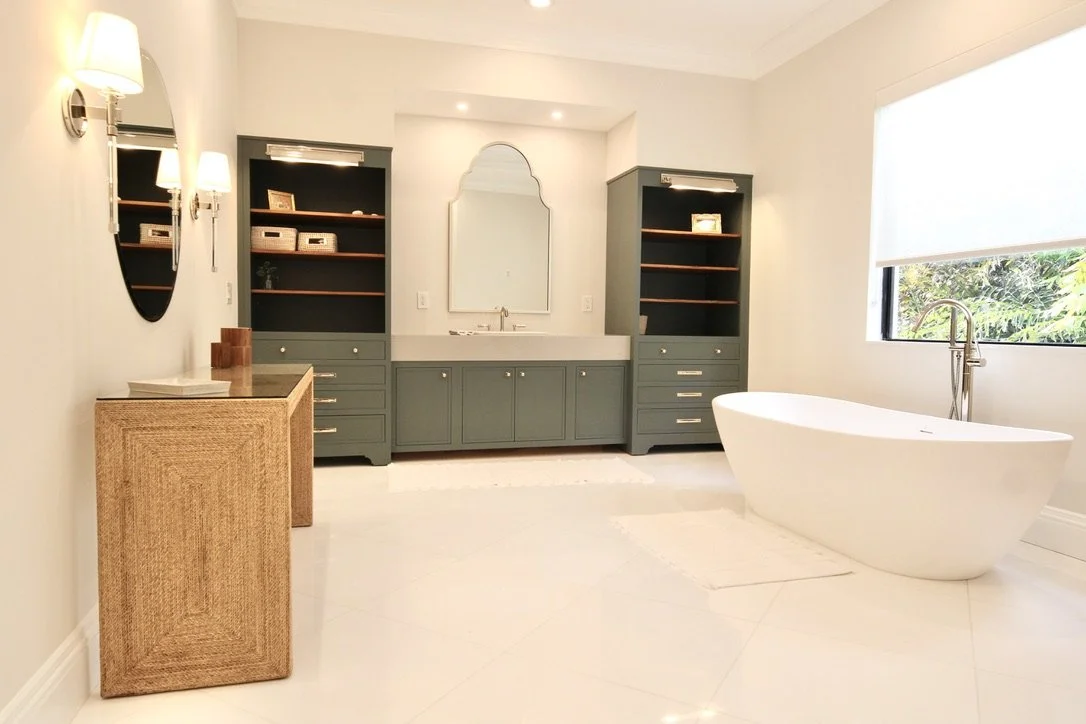 Modern bathroom with a white freestanding bathtub near a large window, a green vanity with a mirror, and two matching green open shelves. A rattan cabinet is on the left, and wall-mounted lamps provide lighting.