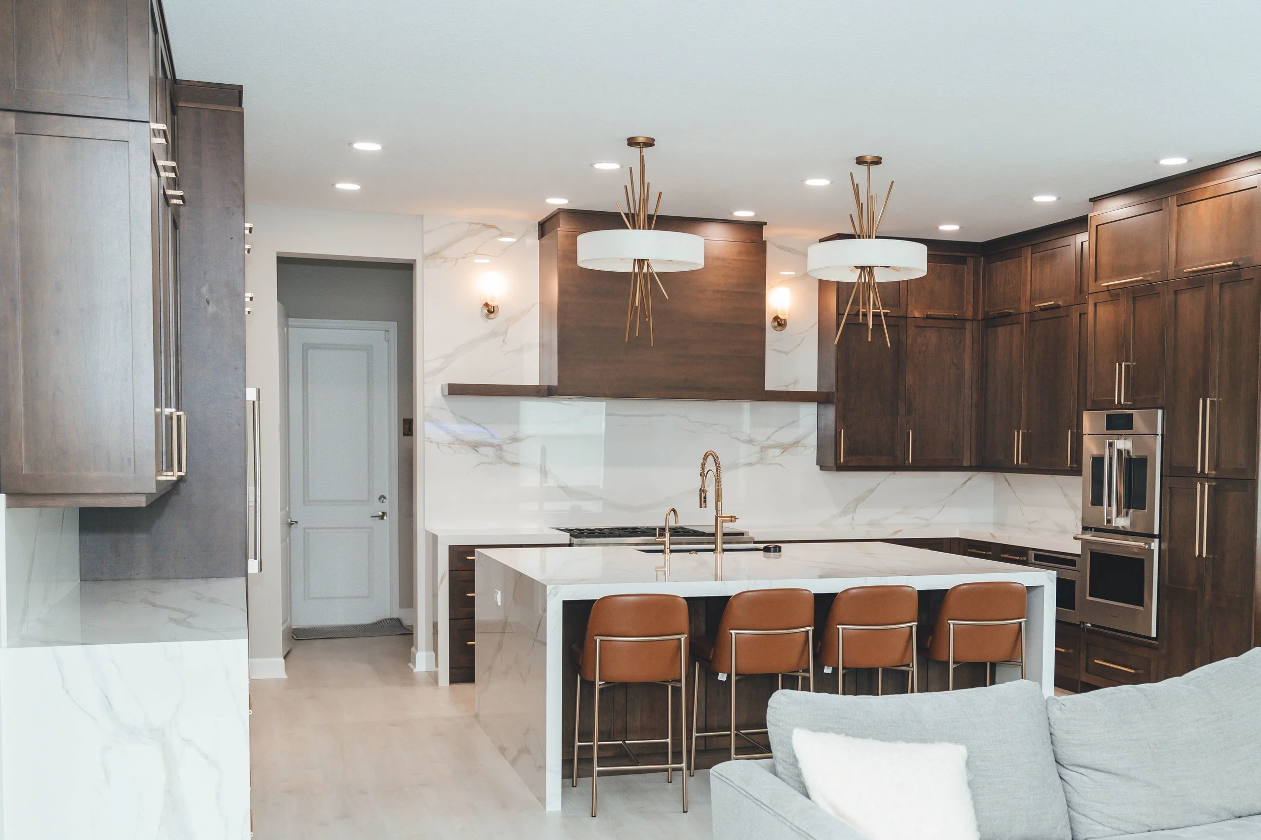 Modern kitchen with white marble countertops, dark wood cabinets, and a kitchen island with four brown chairs. Features include a built-in double oven, a sink with a golden faucet, and decorative modern pendant lights.