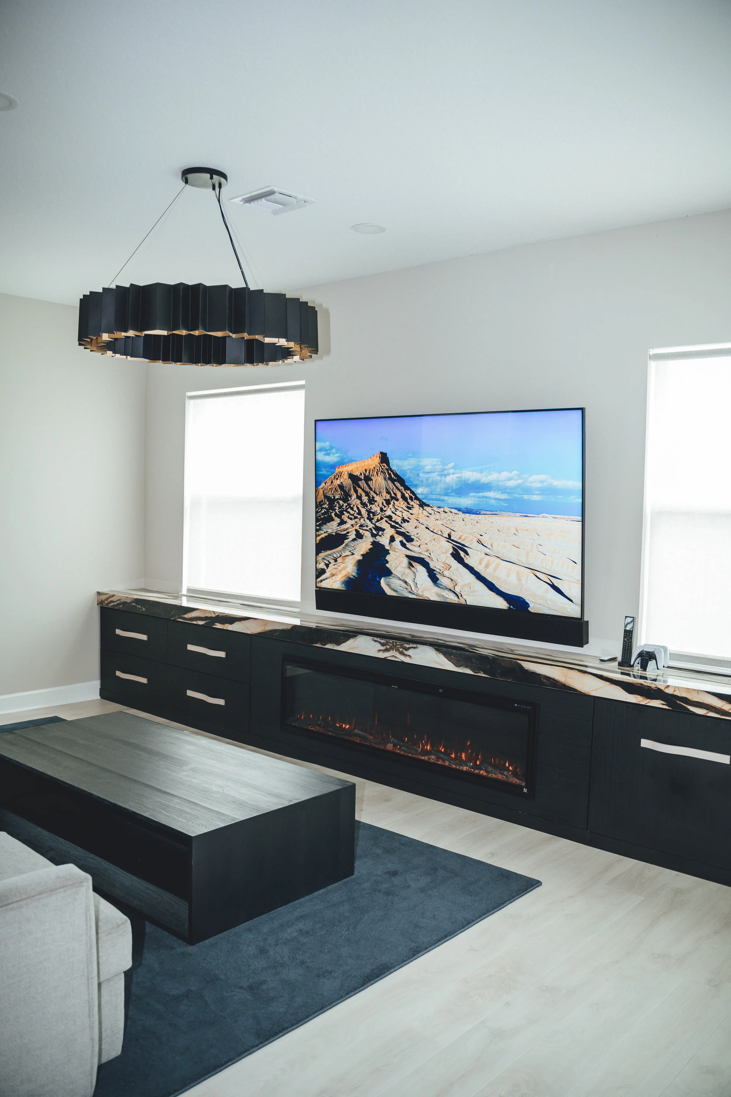Living room with a large flat-screen TV, marble fireplace, black and white sideboard, ceiling light fixture, and windows with blinds.
