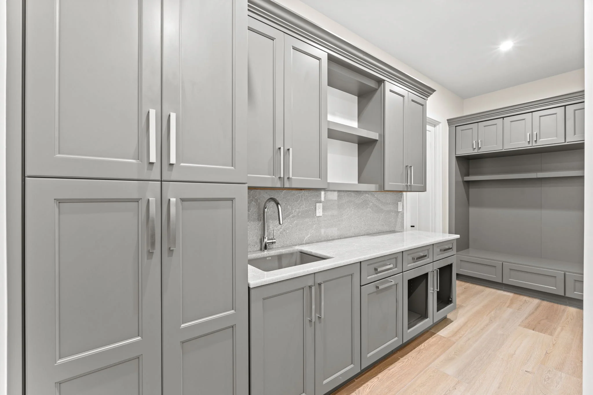 Modern gray kitchen with upper and lower cabinets, a built-in fridge space, a small sink, and open shelving, with light hardwood flooring.
