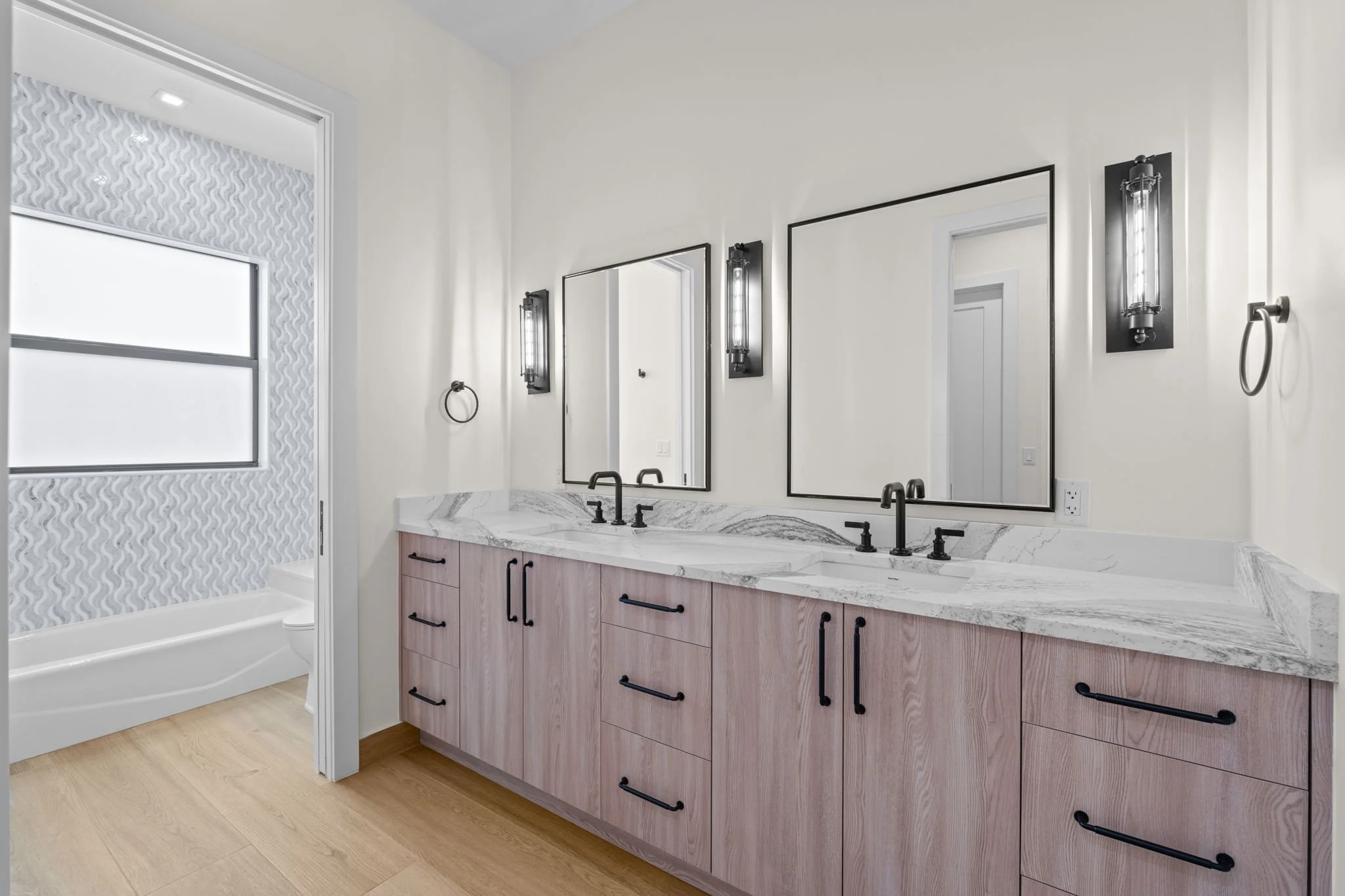 Modern bathroom with a double vanity, white marble countertop, black faucets, large mirrors, black lighting fixtures, white walls, wooden cabinetry, and a separate shower area with geometric patterned tiles.