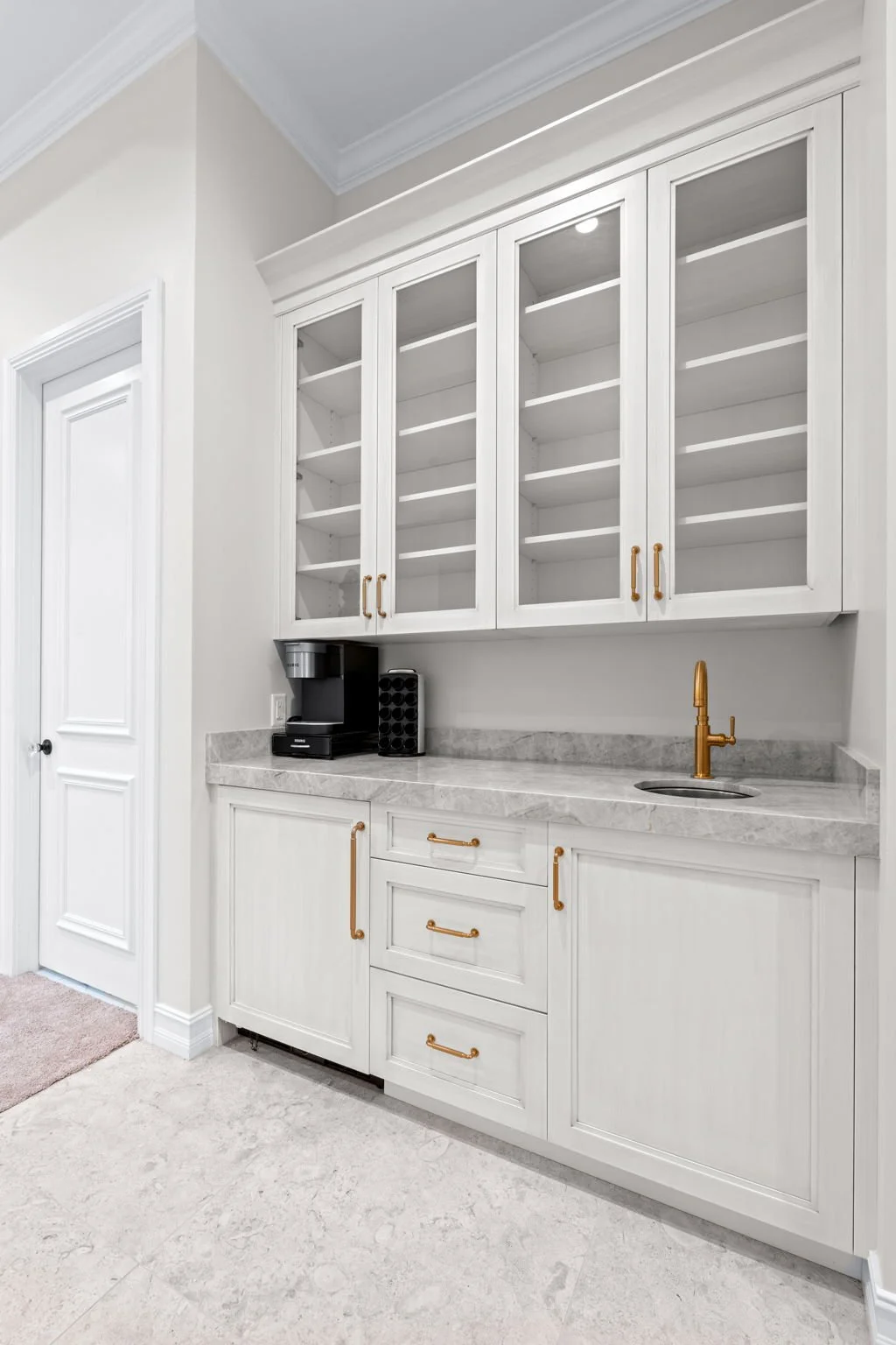 White built-in cabinets with glass doors, gold handles, and a marble countertop in a kitchen or bar area, with a coffee maker and a black spice rack, and a gold faucet over a small sink.