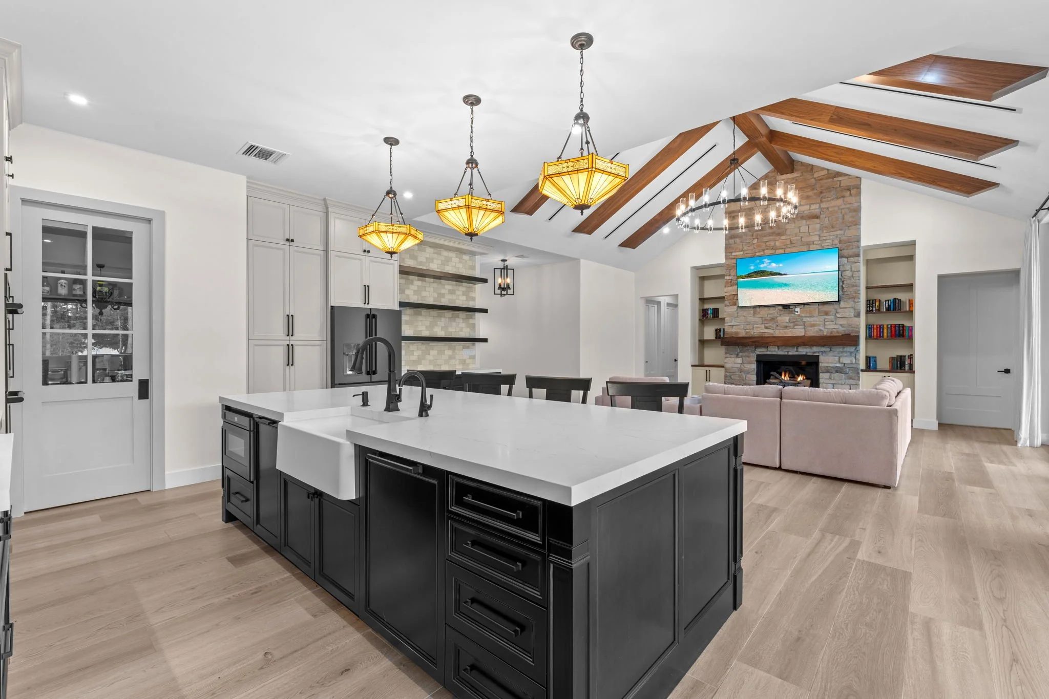 Open-concept living room and kitchen with black island, white countertops, beige flooring, and a stone fireplace with a wall-mounted TV.