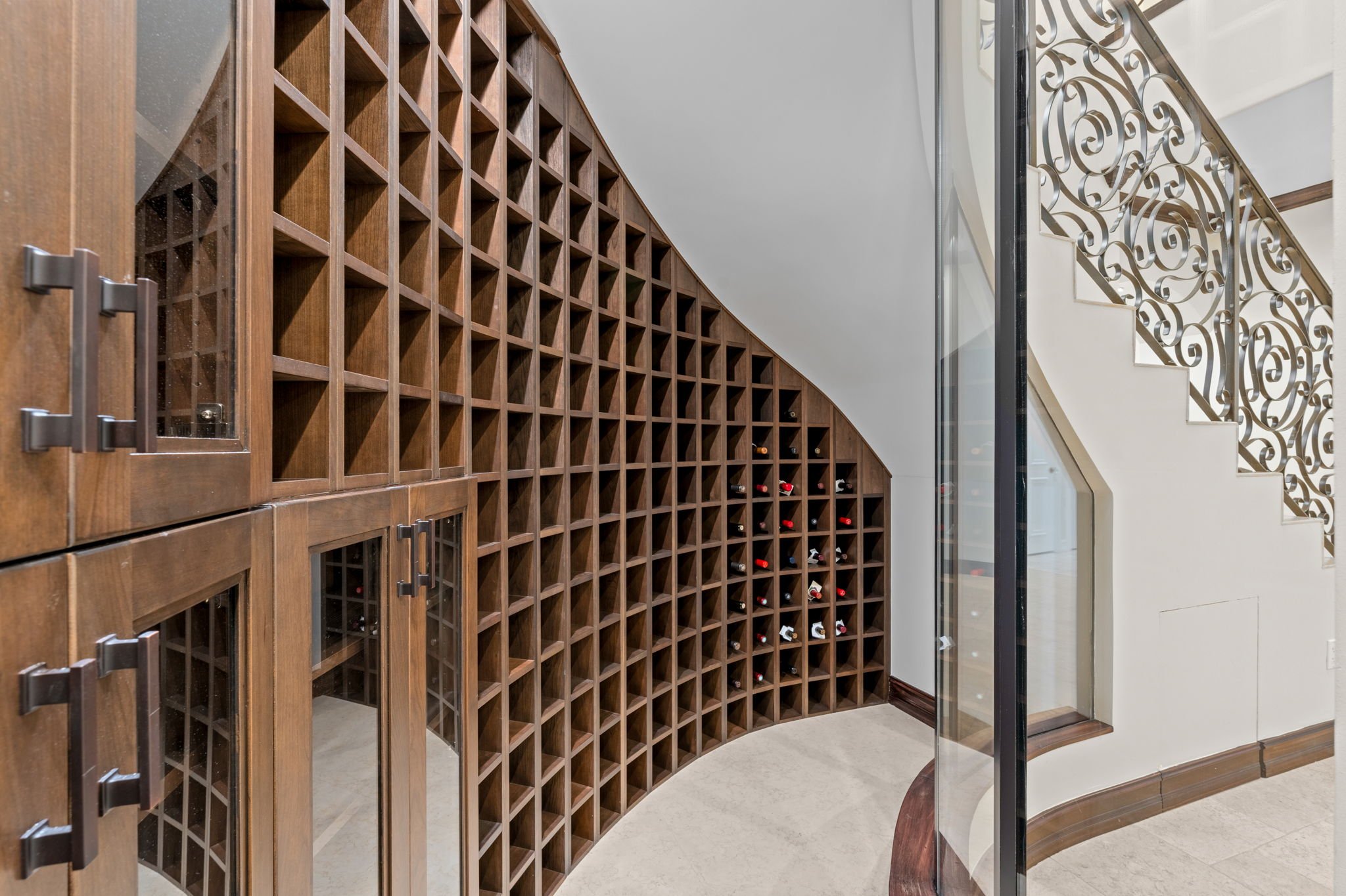 A curved wooden wine rack filled with various bottles in a home interior next to a staircase with ornate ironwork railing.