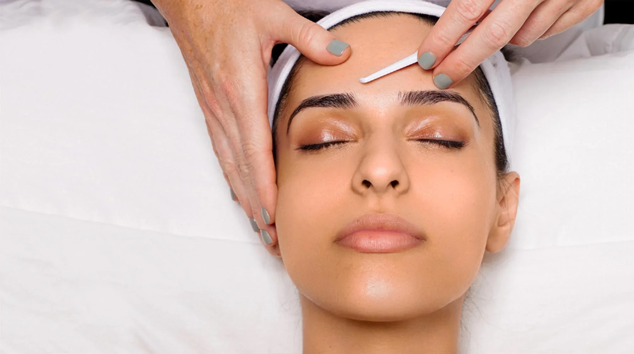 A woman with closed eyes receives a facial treatment while lying on a white bed. A person with painted nails is using a cotton swab on her forehead.