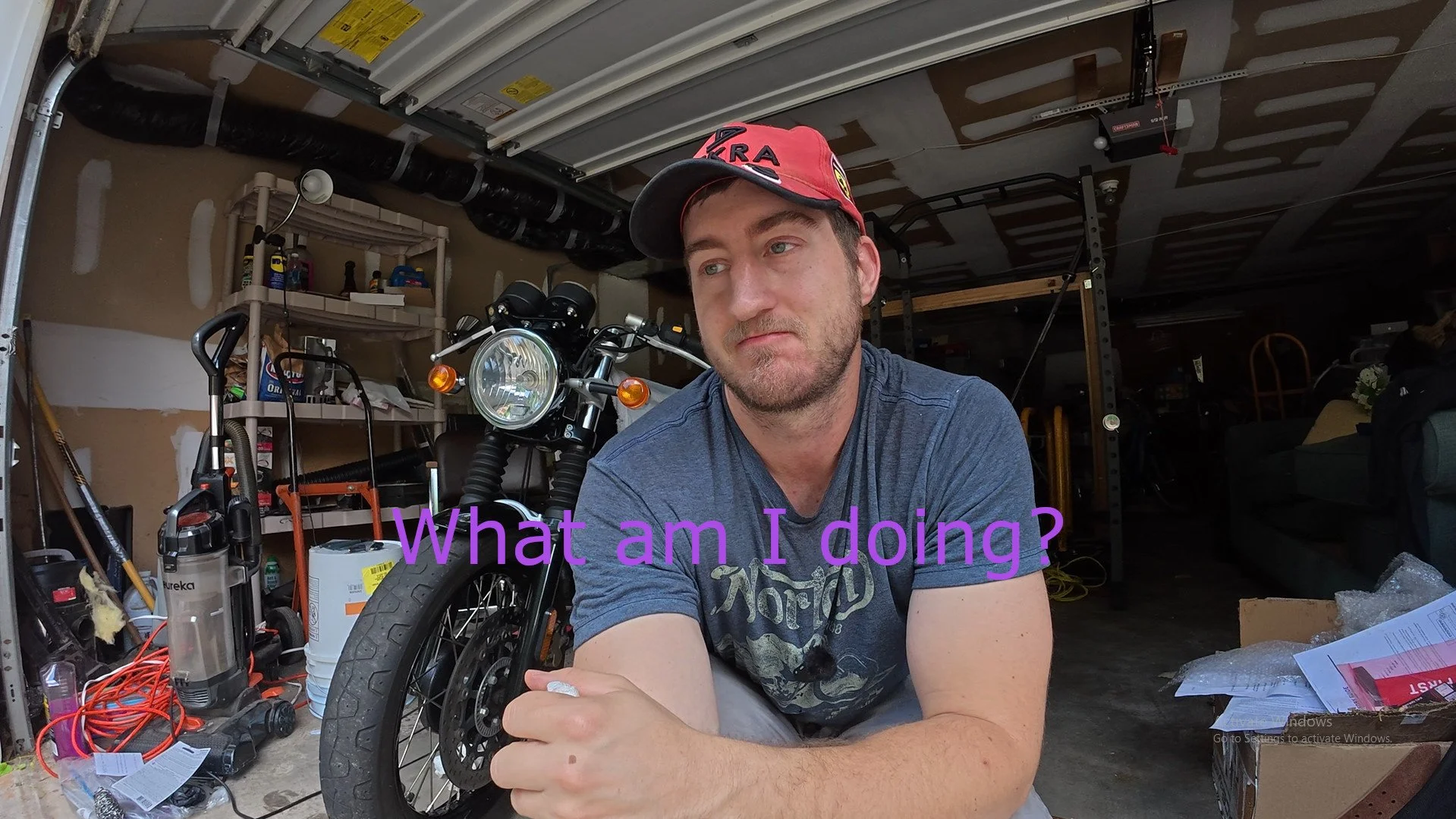 A man with a beard and a gray T-shirt, wearing a red cap, sitting in a garage next to a motorcycle, with tools and clutter in the background. Purple text overlaid on the image says, 'What am I doing?'.