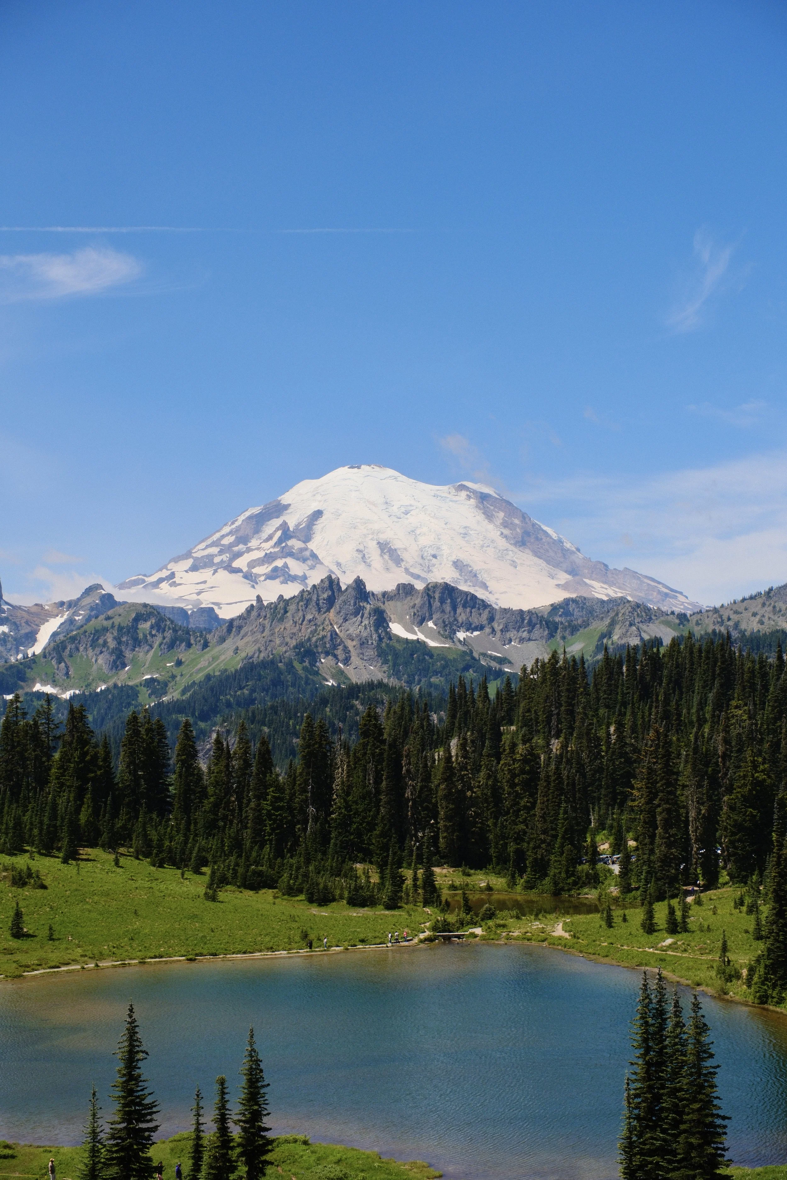 Snow-capped mountain surrounded by green forest and a small lake in the foreground, under a clear blue sky.