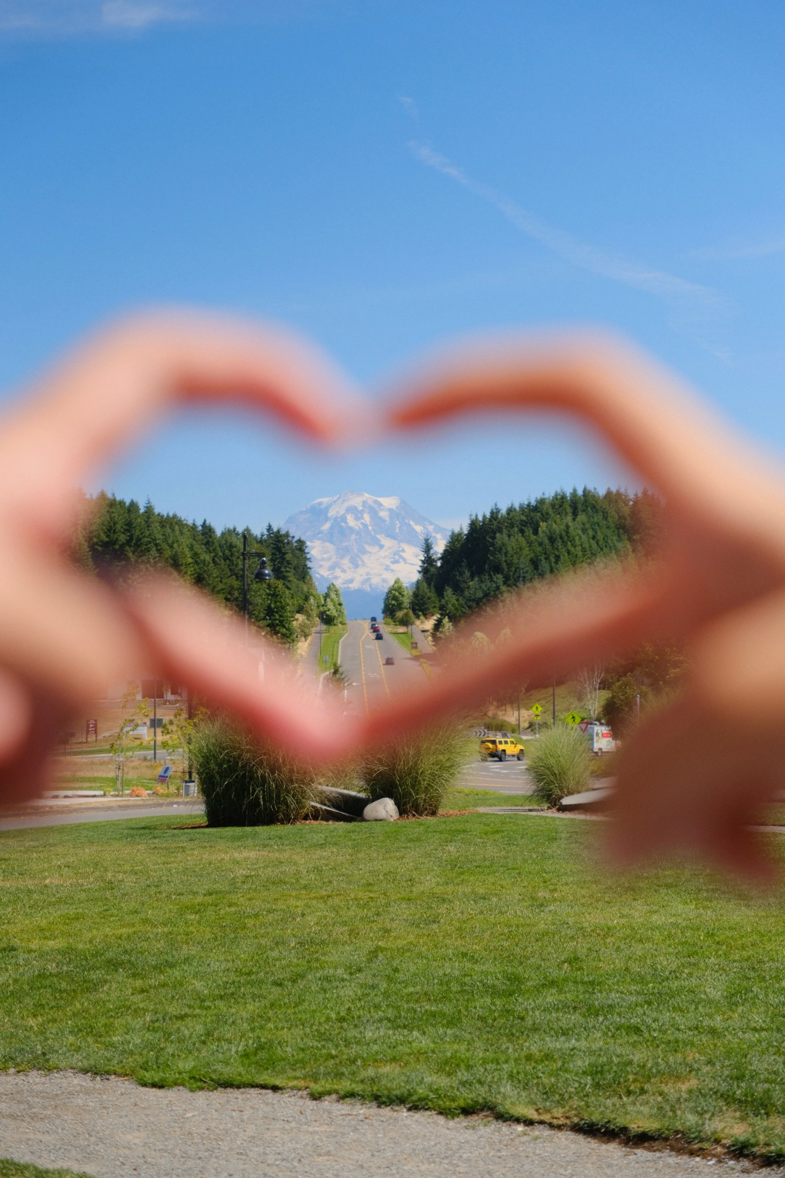 Person making a heart shape with their hands, framing a distant snowy mountain under a blue sky with clouds.