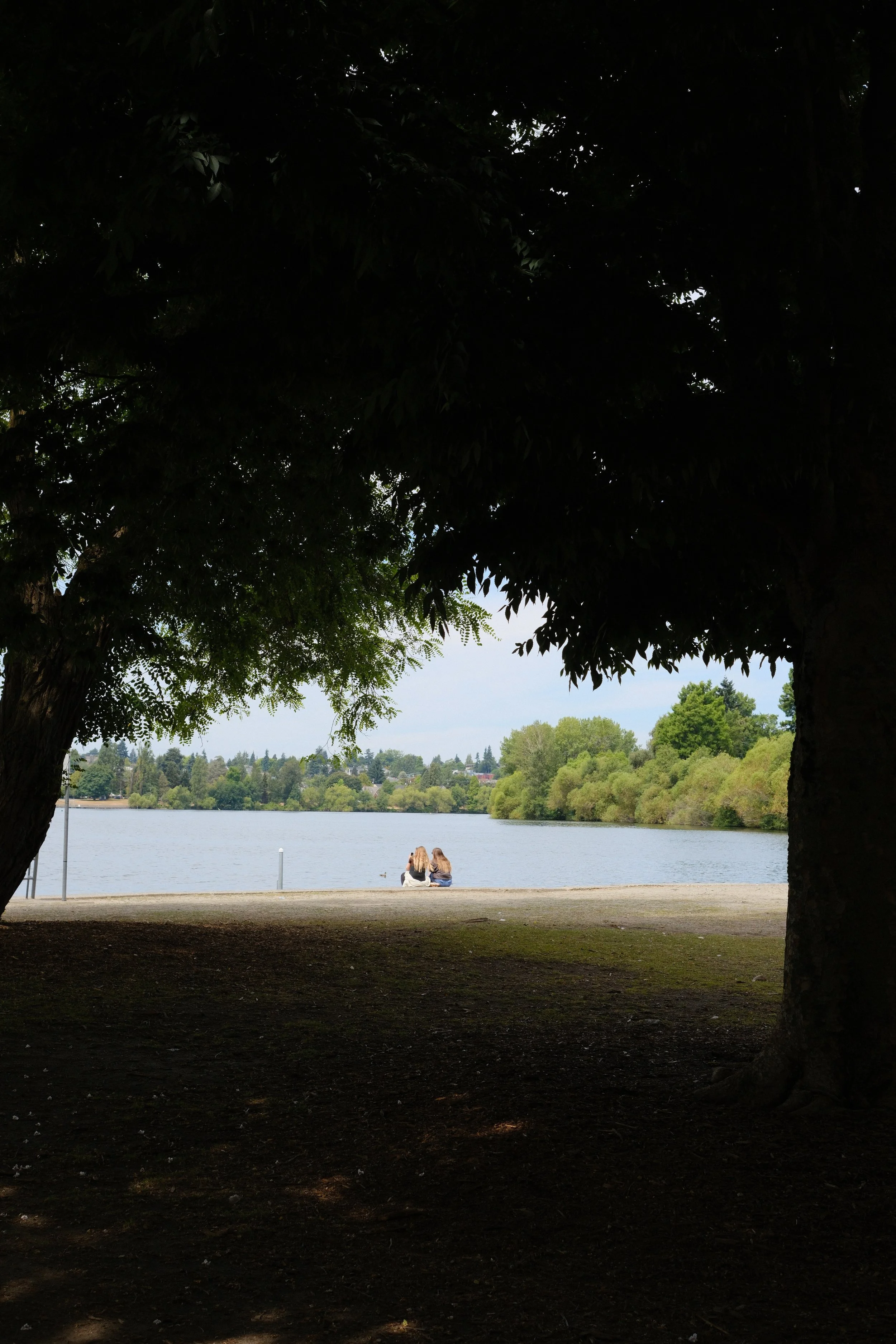 Two women sitting by a lake, seen through the shade of large trees.