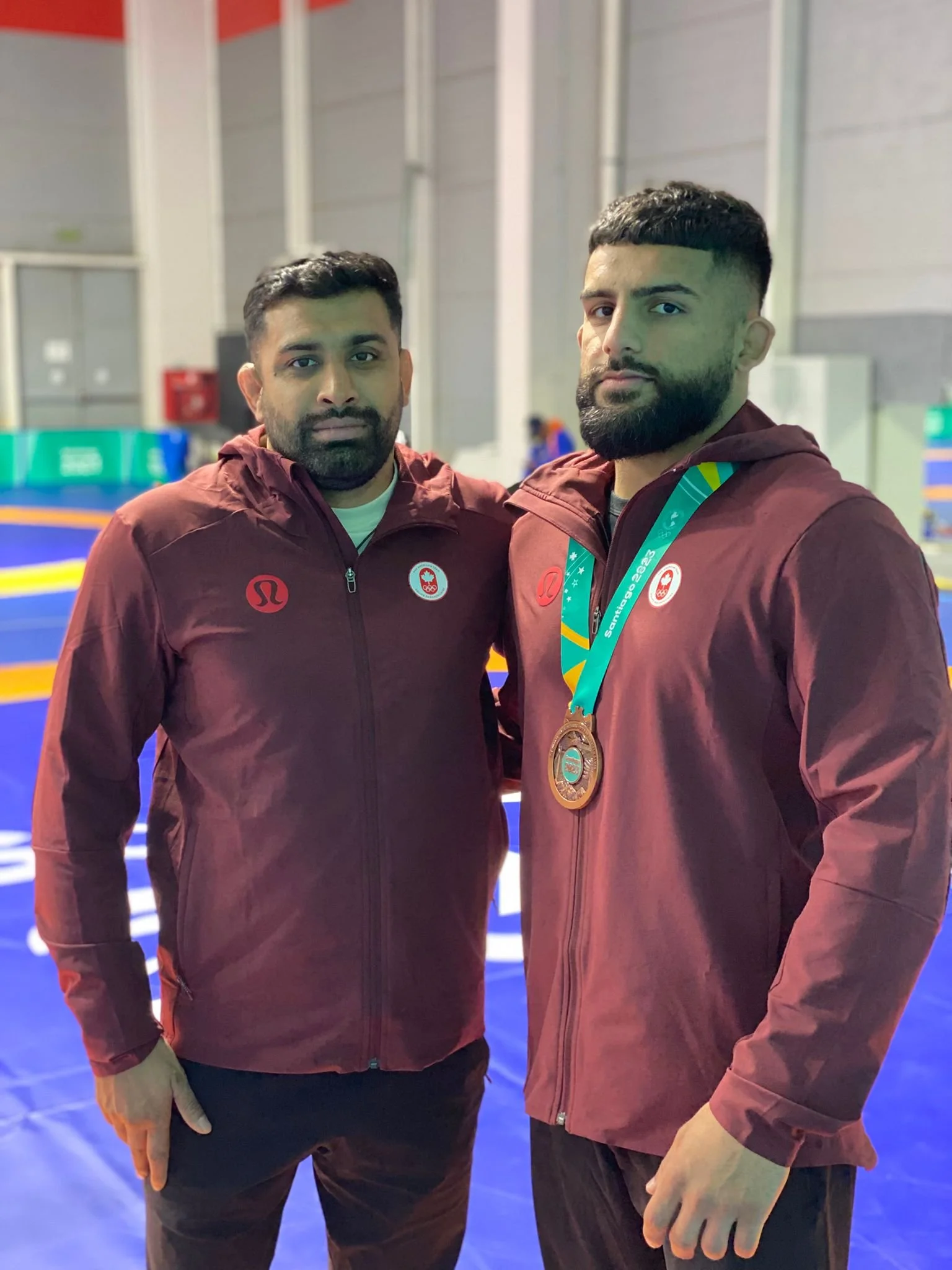 Two male athletes wearing maroon jackets with logos, one with a gold medal around his neck, standing inside a sports facility with a blue mat background.