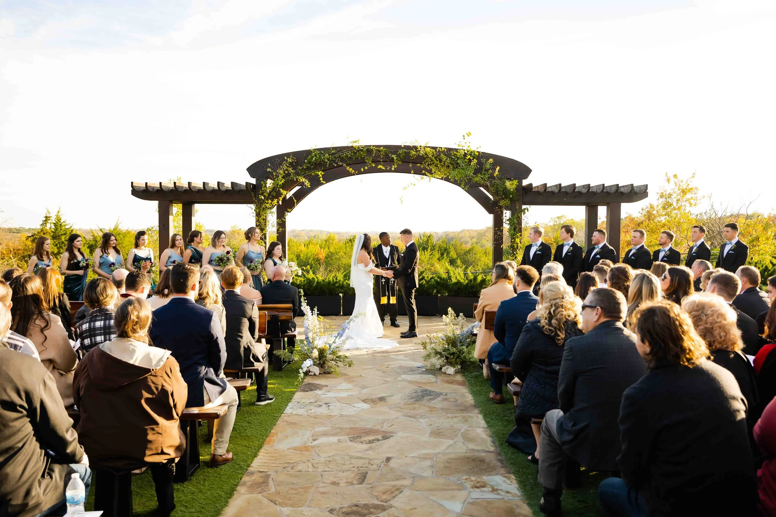 Outdoor wedding ceremony with a bride and groom exchanging vows under a wooden arch, surrounded by bridesmaids and groomsmen, with guests seated on either side.