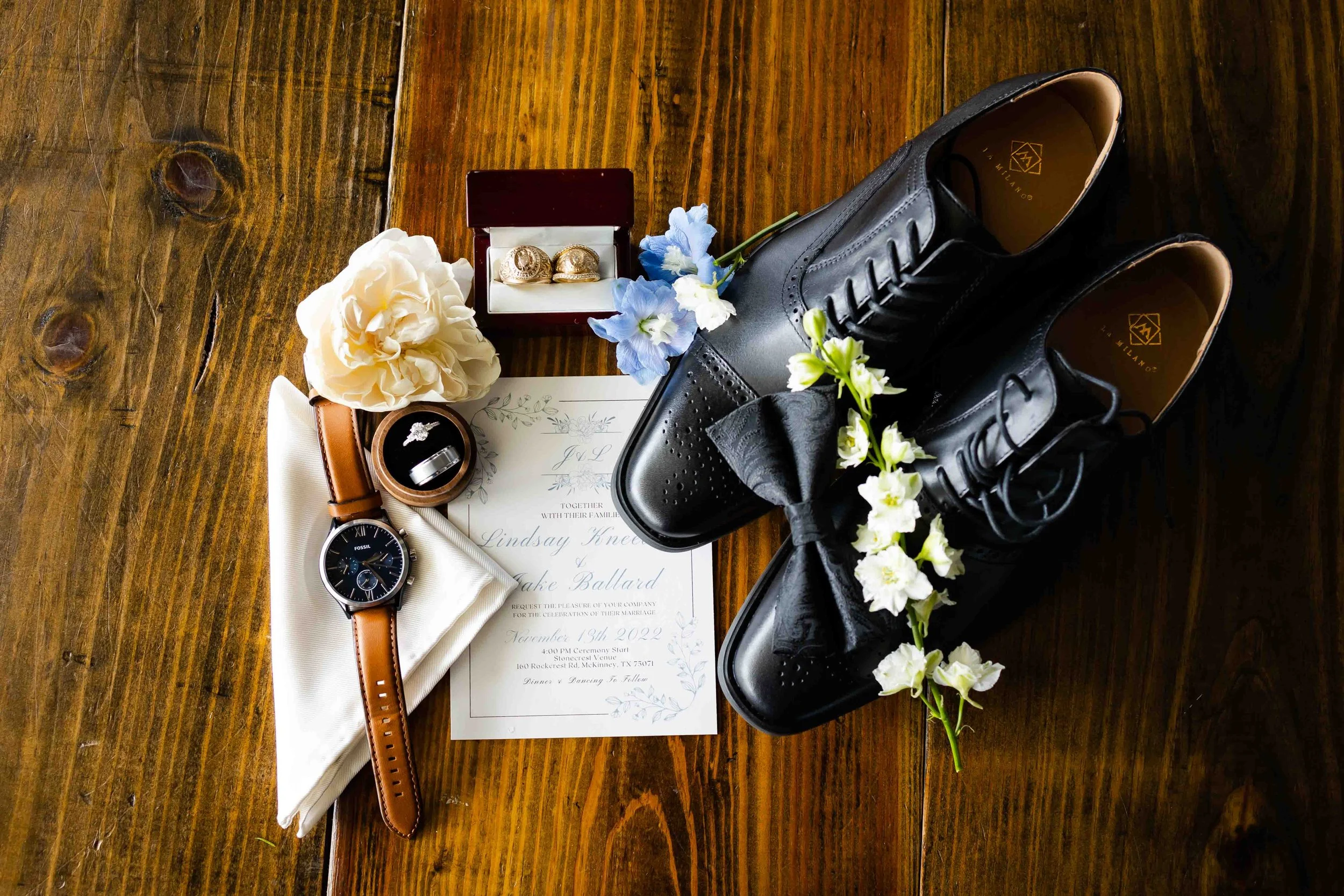 Wedding shoes, rings, watch, flowers, invitation, and jewelry on a wooden surface.
